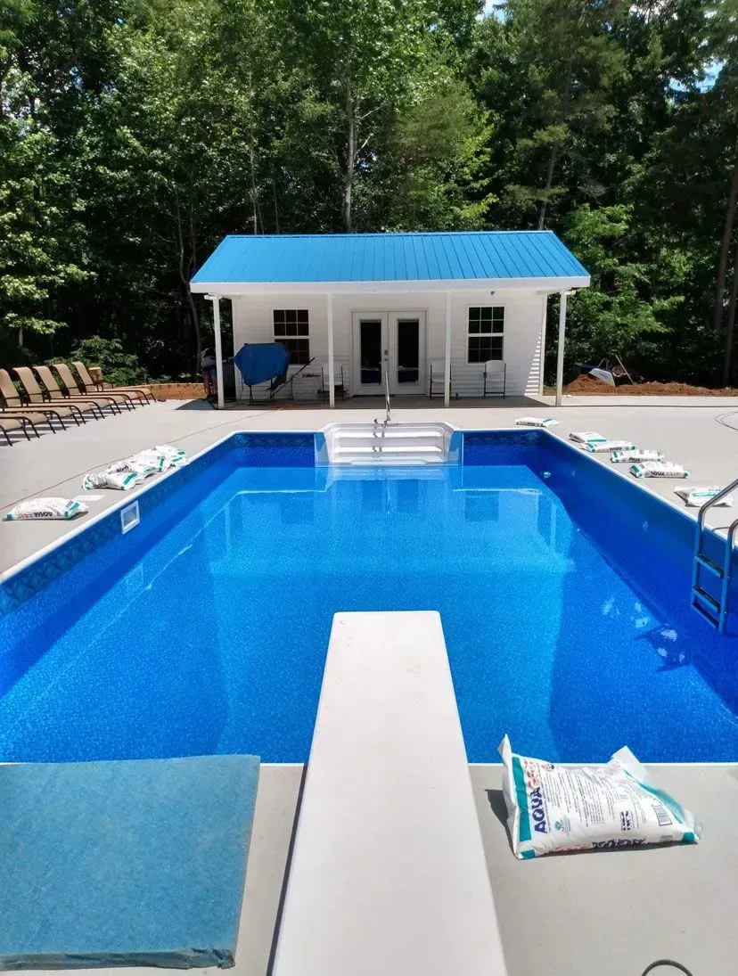 A rectangular blue swimming pool with a diving board, surrounded by a concrete patio and a small white building with a blue roof, trees in the background.