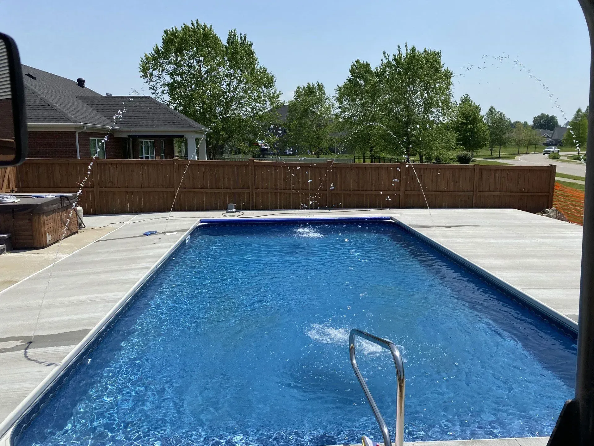 Rectangular swimming pool with blue water and fountain jets, concrete patio, brown fence, and green trees under a blue sky.
