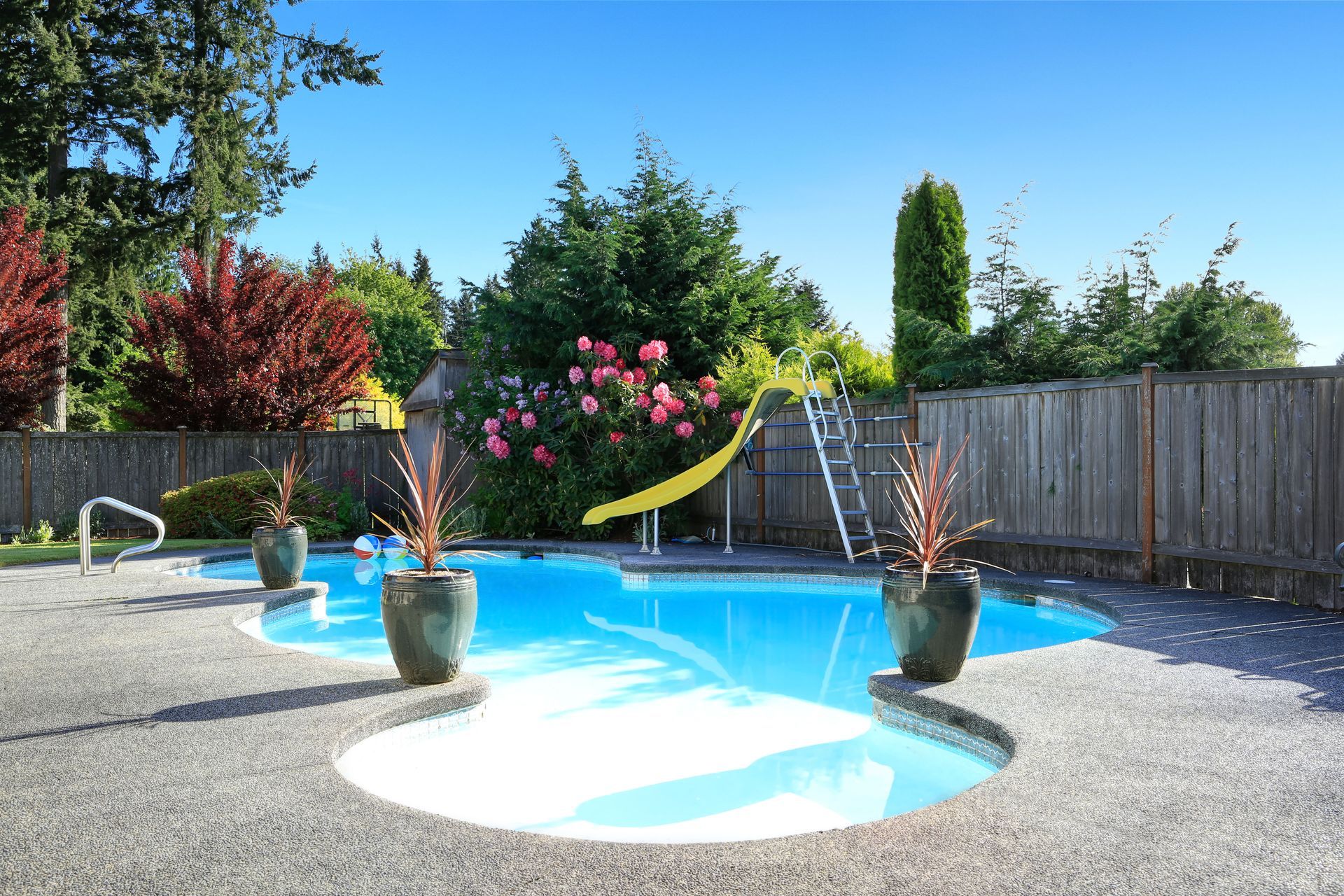 A bright blue backyard swimming pool with a yellow slide, surrounded by a gravel patio, green trees, and a wooden fence.