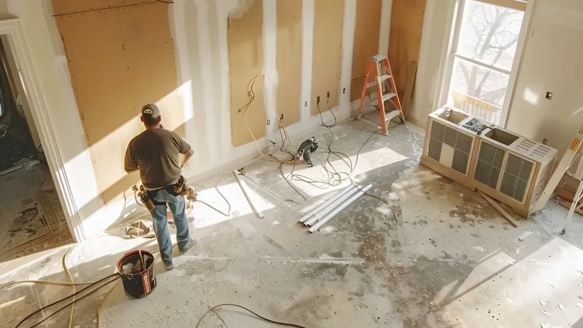 A worker stands in a room undergoing renovation, featuring unfinished walls, a stepladder, tools, and construction debris.