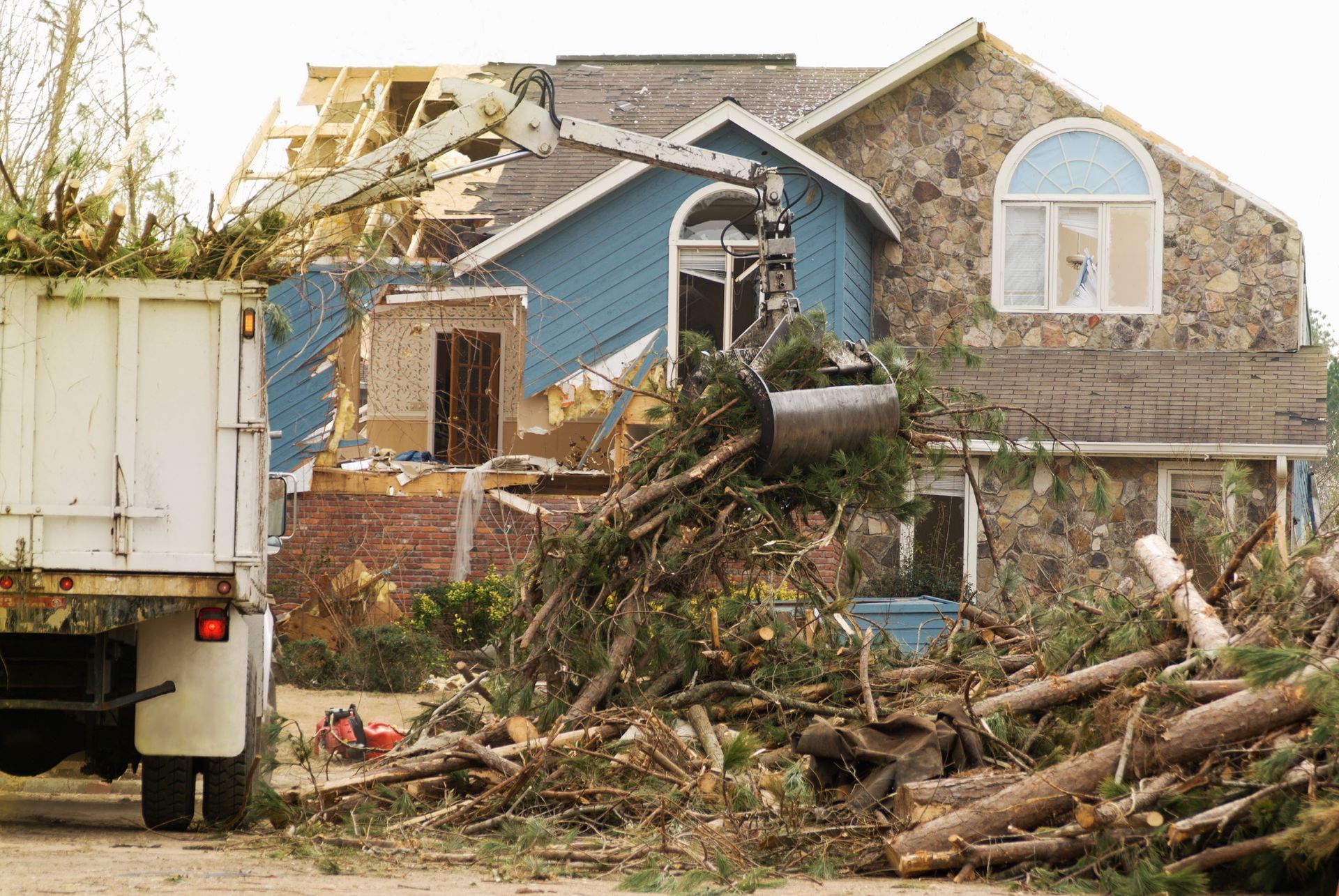 A loader arm collects storm debris in front of a house with significant structural damage and broken windows.