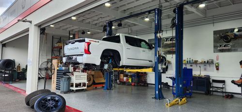 White truck on a lift inside a garage. Other car parts are on the floor.