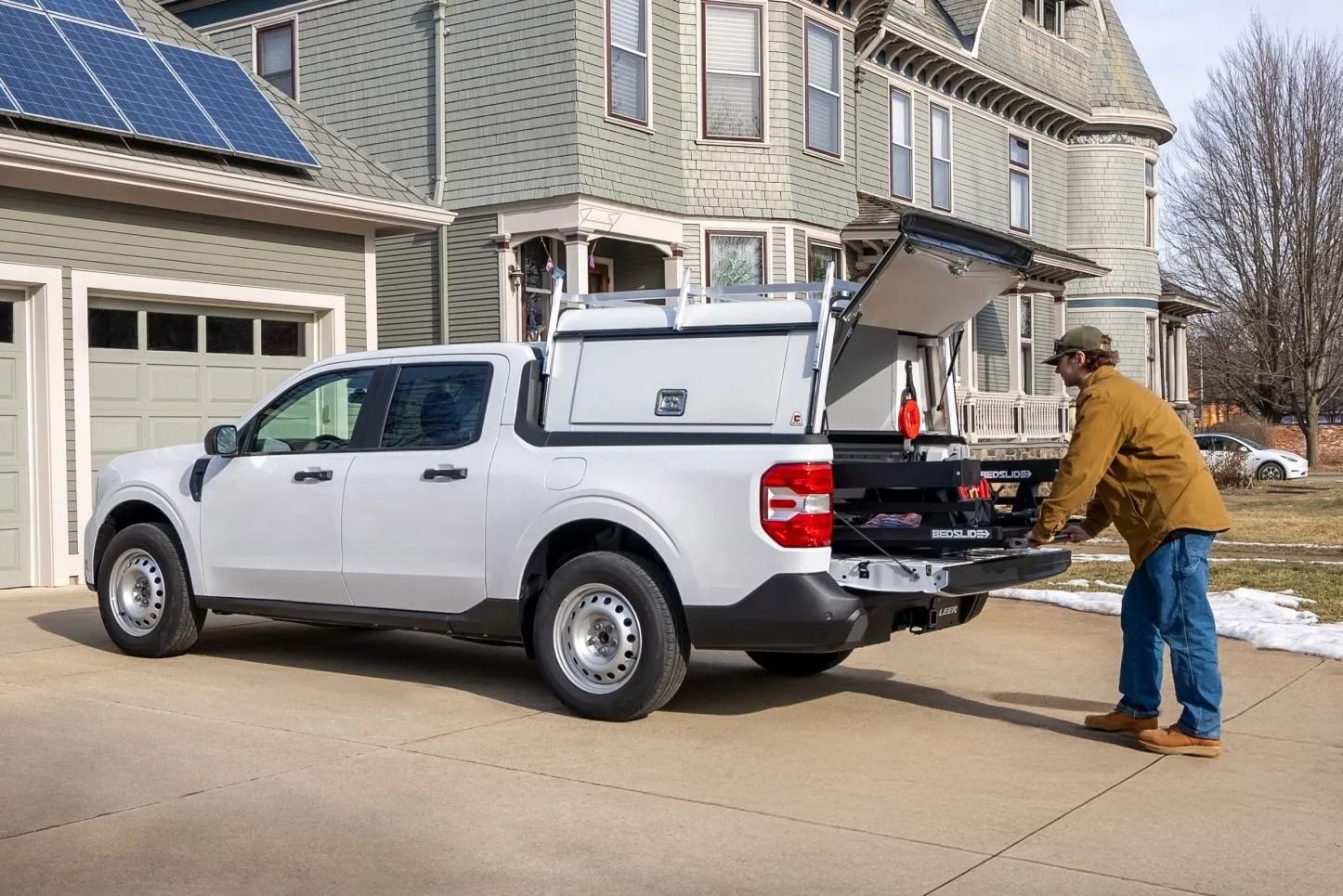 White pickup truck with utility bed, man reaching in. Garage and house in the background. Sunny day.