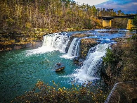 A waterfall in the middle of a river with a bridge in the background.