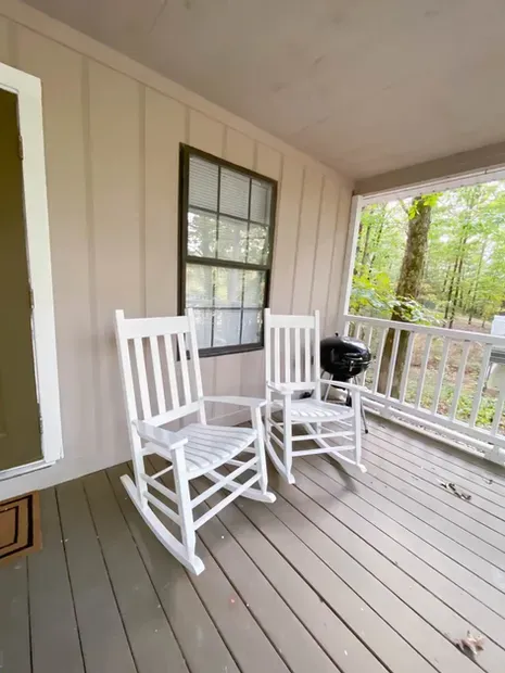 Two white rocking chairs are sitting on a porch next to a window.