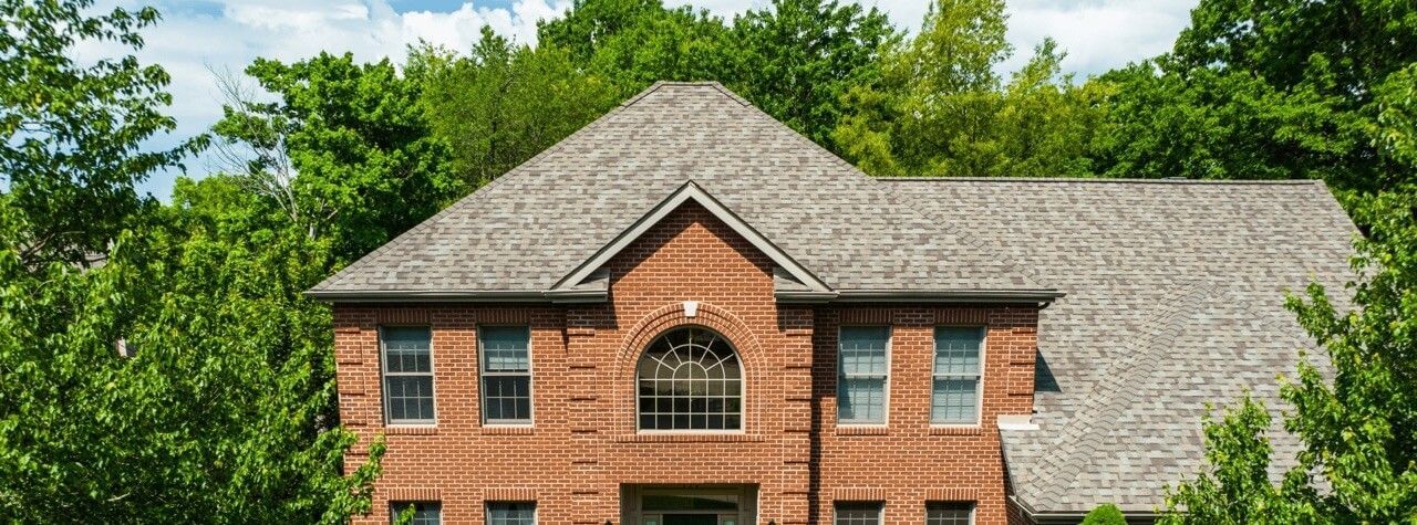 A large brick house with a gray roof is surrounded by trees.