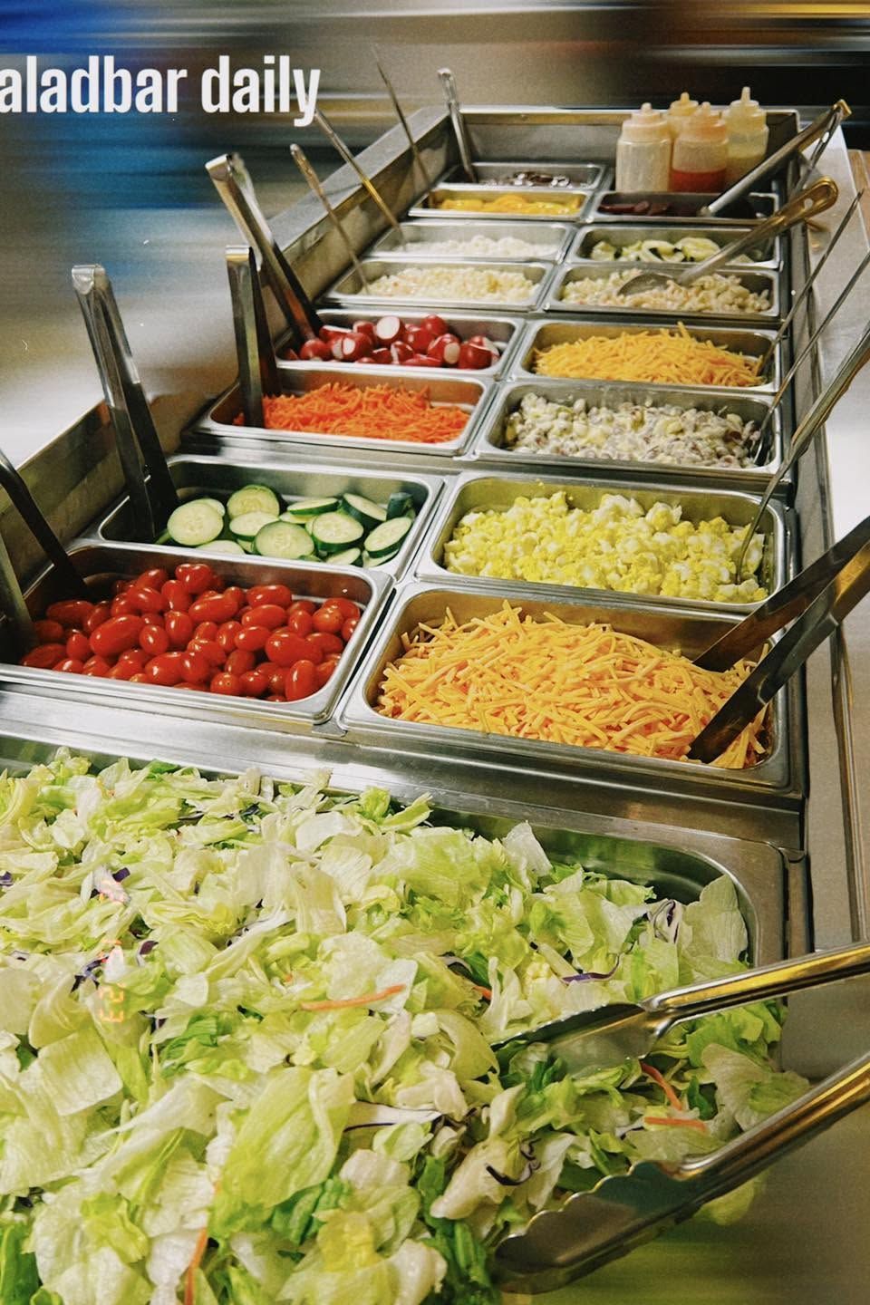 Salad bar with trays of lettuce, tomatoes, cucumbers, and assorted toppings in a restaurant setting