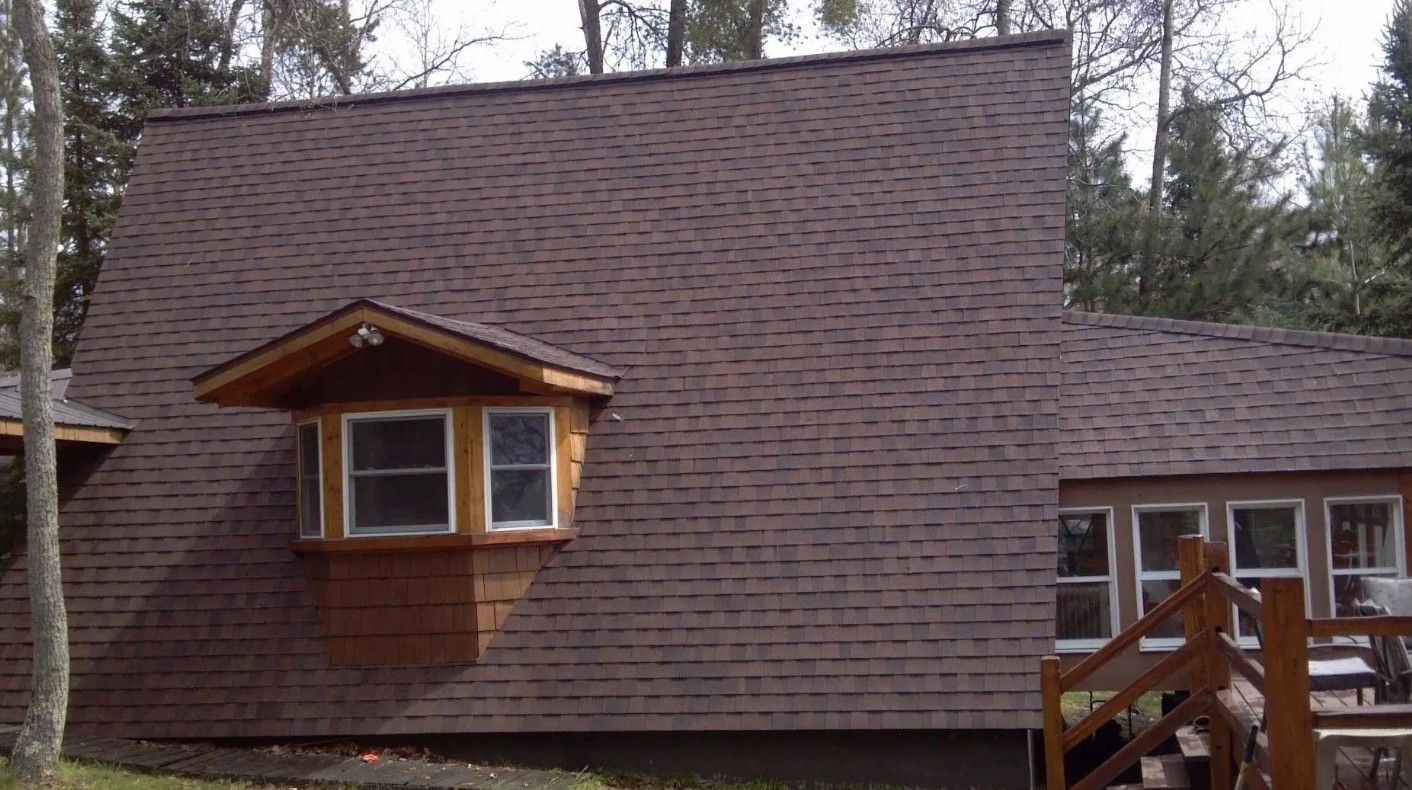 Brown shingled cabin roof with dormer windows amid trees and a wooden deck