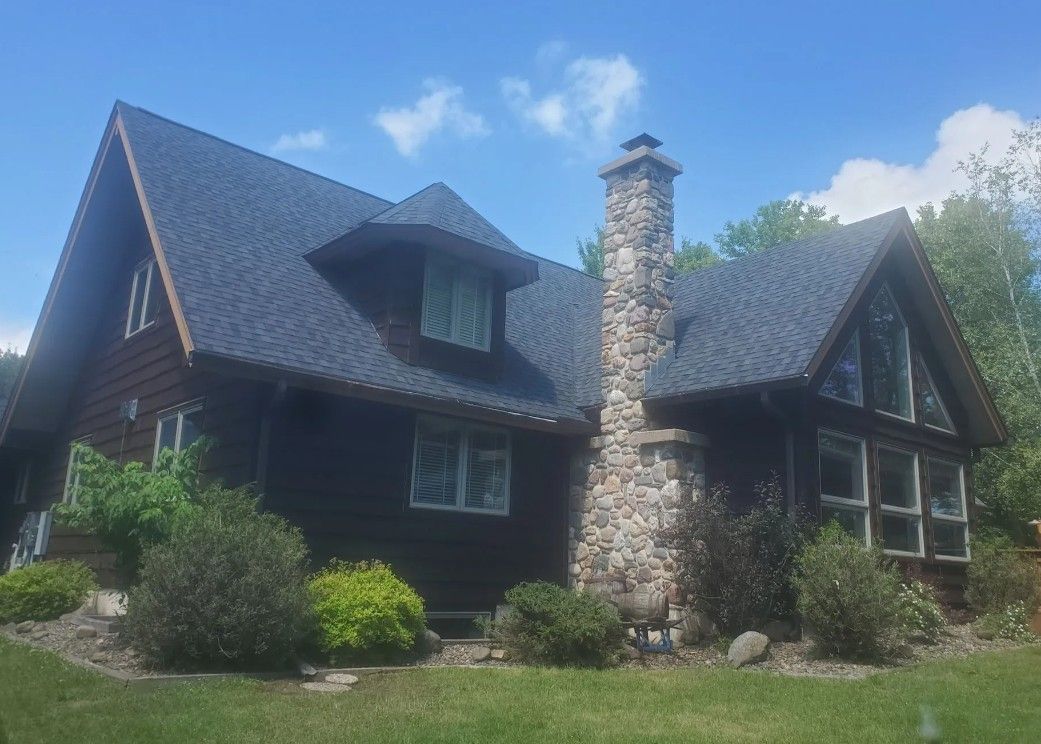 Log cabin-style house with a stone chimney and dark roof on a sunny, grassy lot