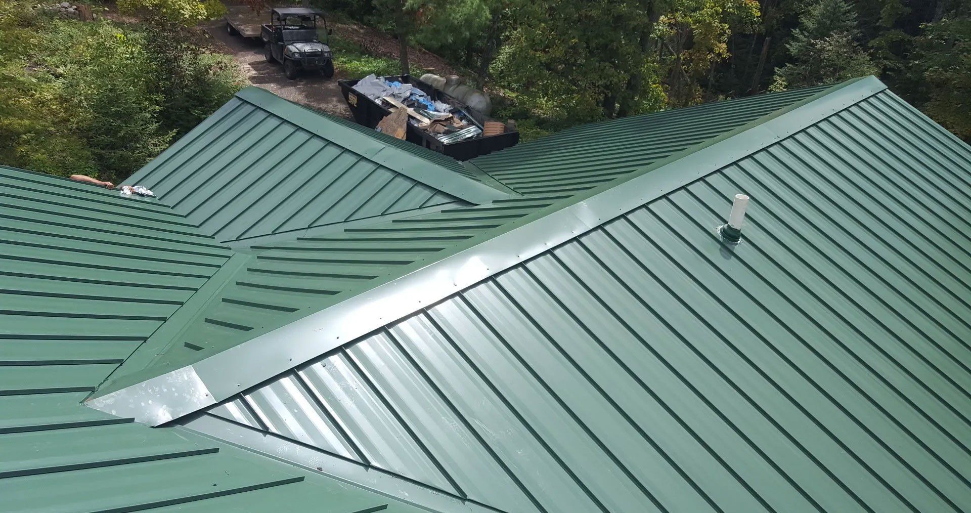 Green metal roof with a silver ridge vent, viewed from above, with trees and parked vehicles in the background