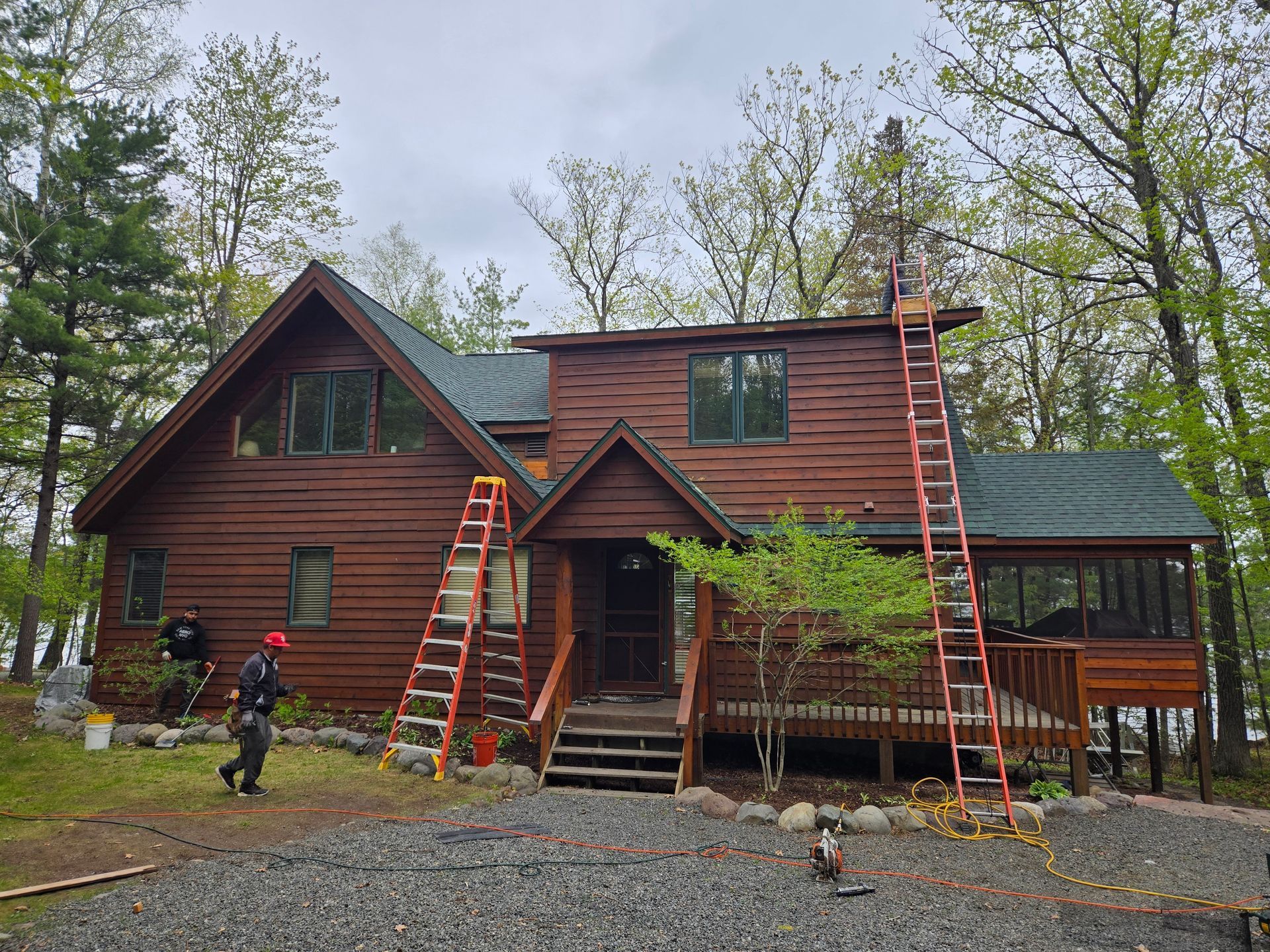 Two-story brown cabin with ladders and workers on the roof, surrounded by trees.