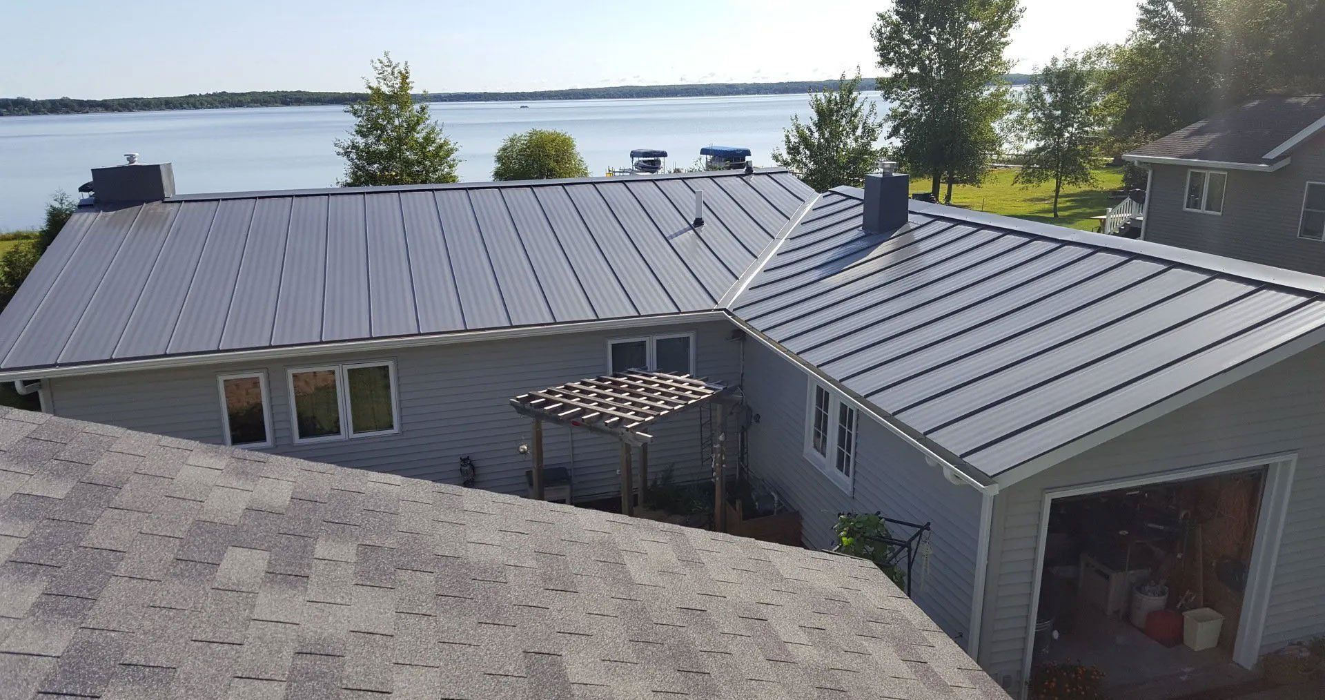 Rooftops of a lakeside house with gray metal roofs, skylights, and a view of water and trees beyond.
