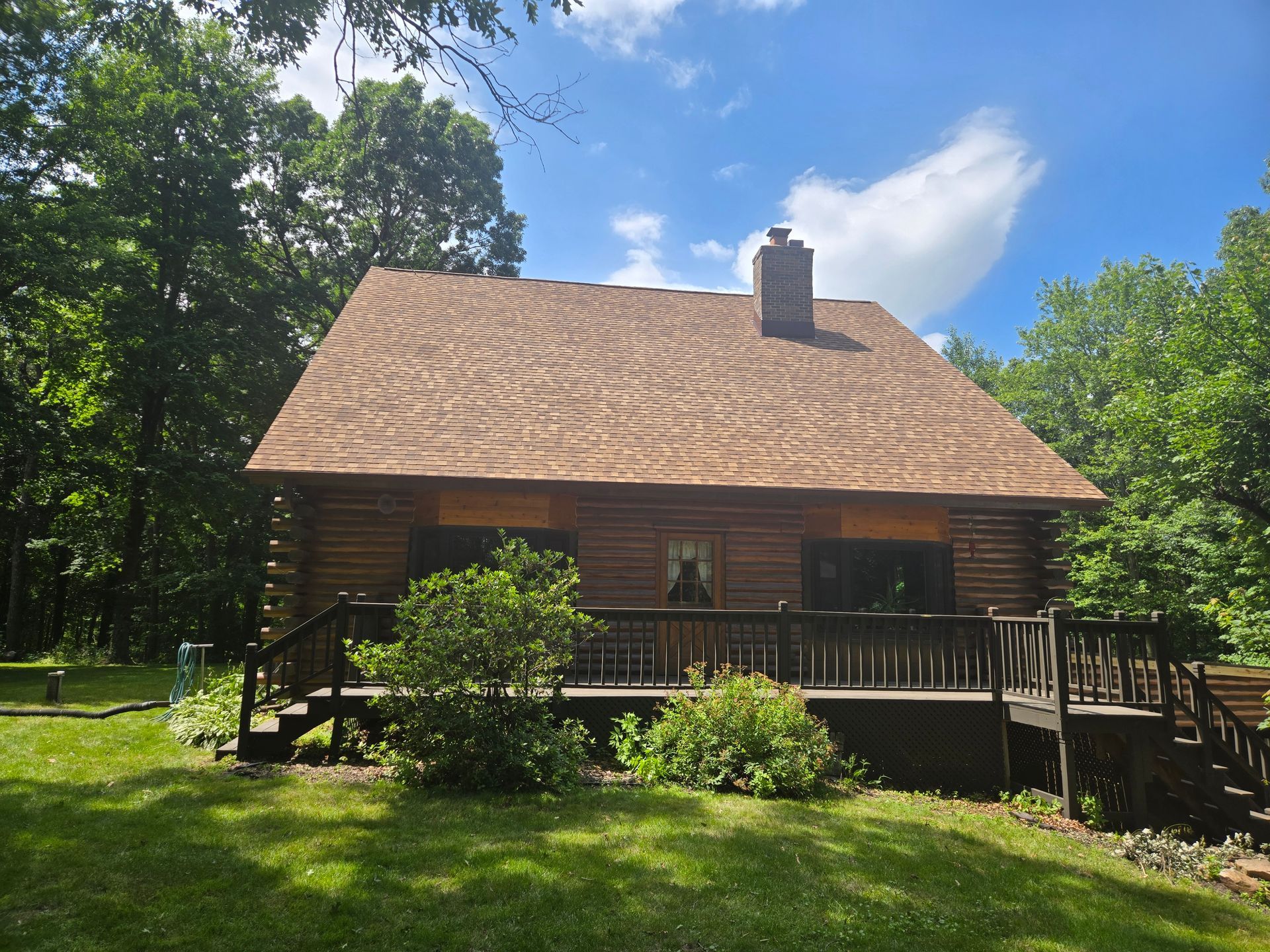 Rustic wooden cabin with a steep roof and chimney, surrounded by green trees and lawn under a blue sky