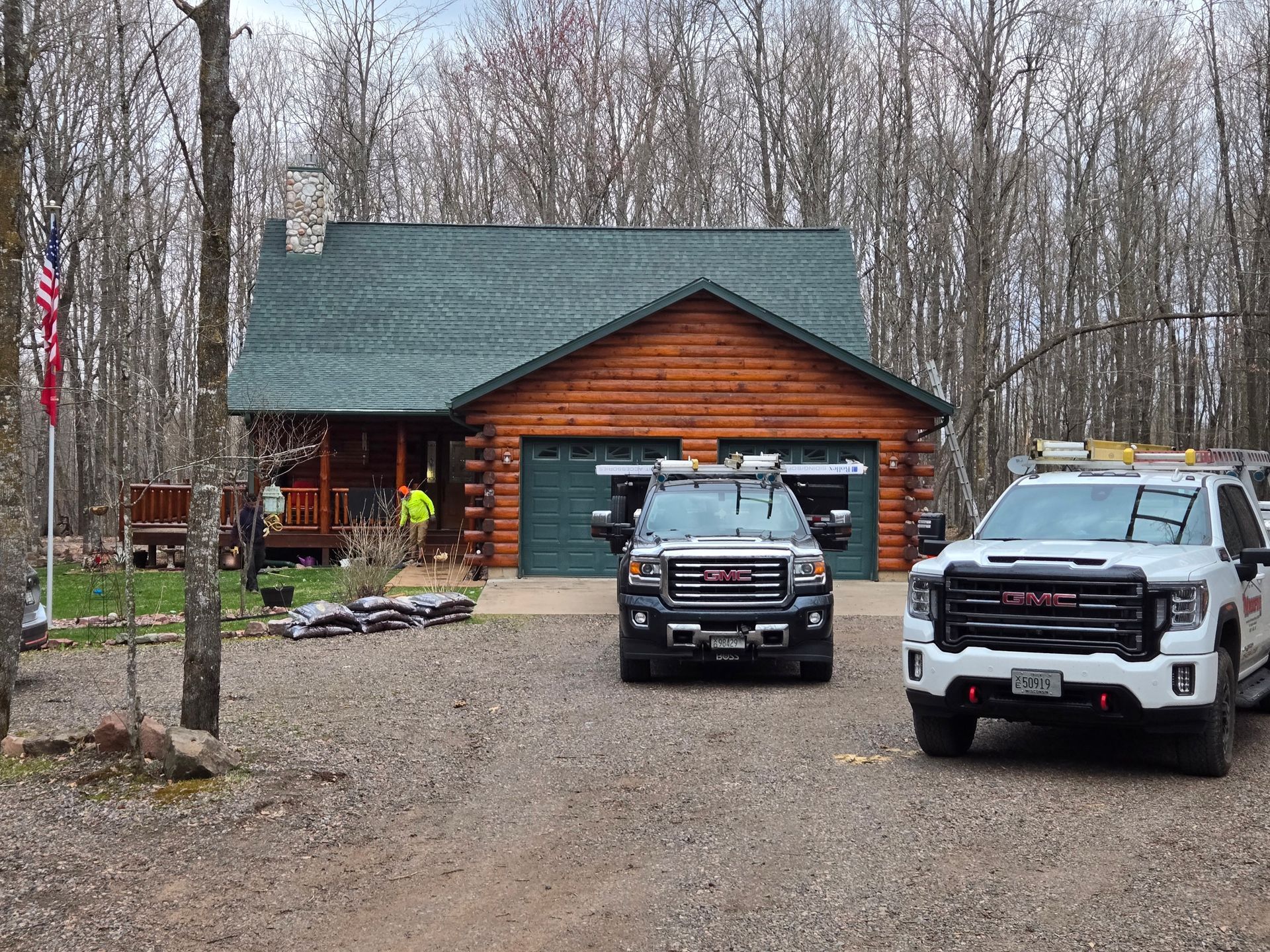 Log cabin home with green roof, two trucks in gravel driveway, and bare trees in the background