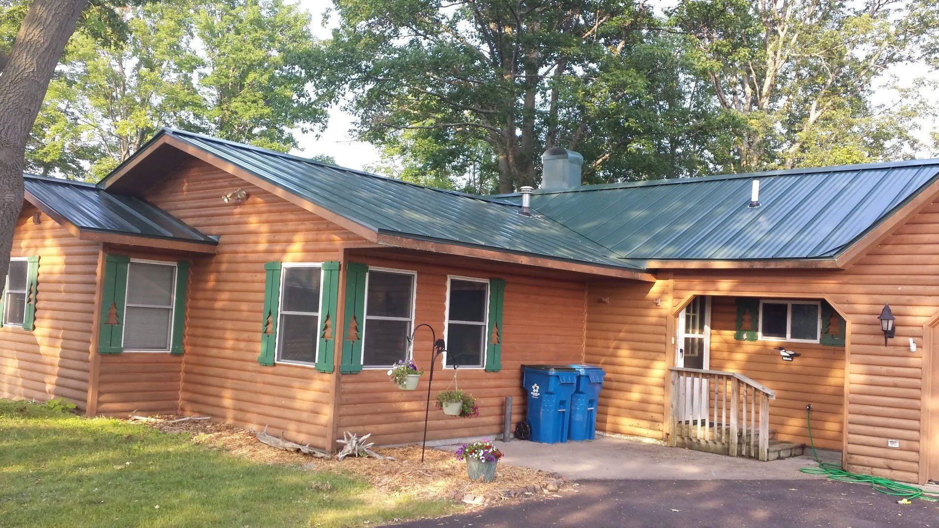 Wooden cabin-style house with green-trimmed windows, blue bins, and a small porch under trees