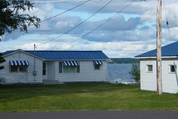 White lakeside cabins with blue roofs beside a grassy shore and water under cloudy skies