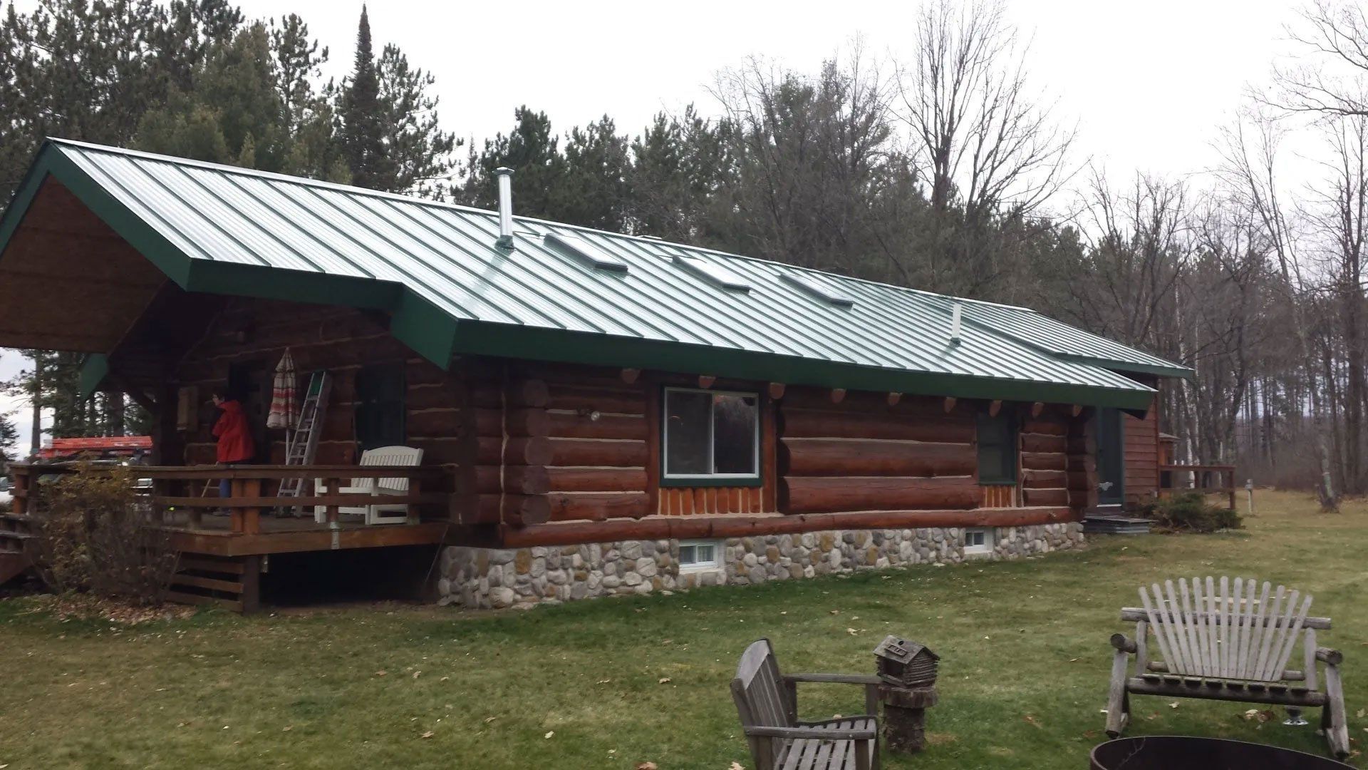 Log cabin with green metal roof on a grassy lot, with chairs in front and trees behind.