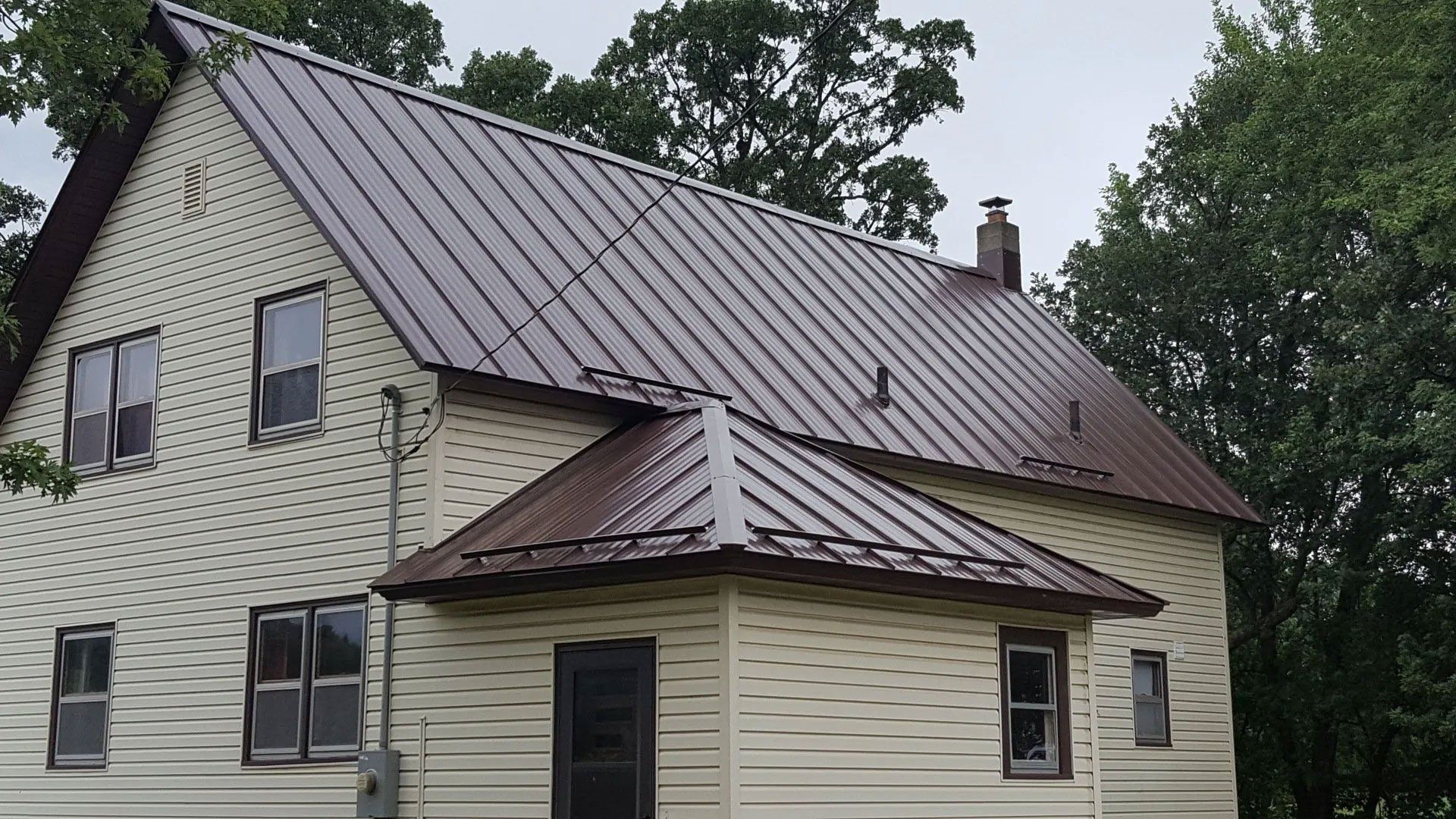 Two-story cream house with a brown metal roof and trees in the background