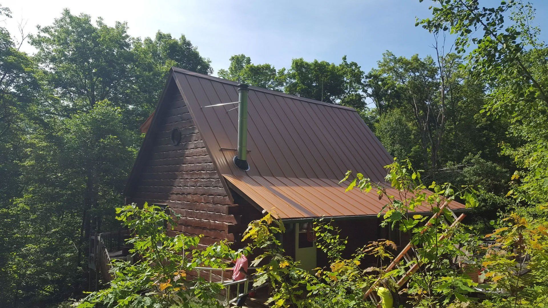 Small wooden cabin with a brown roof nestled among green trees and shrubs under a blue sky.