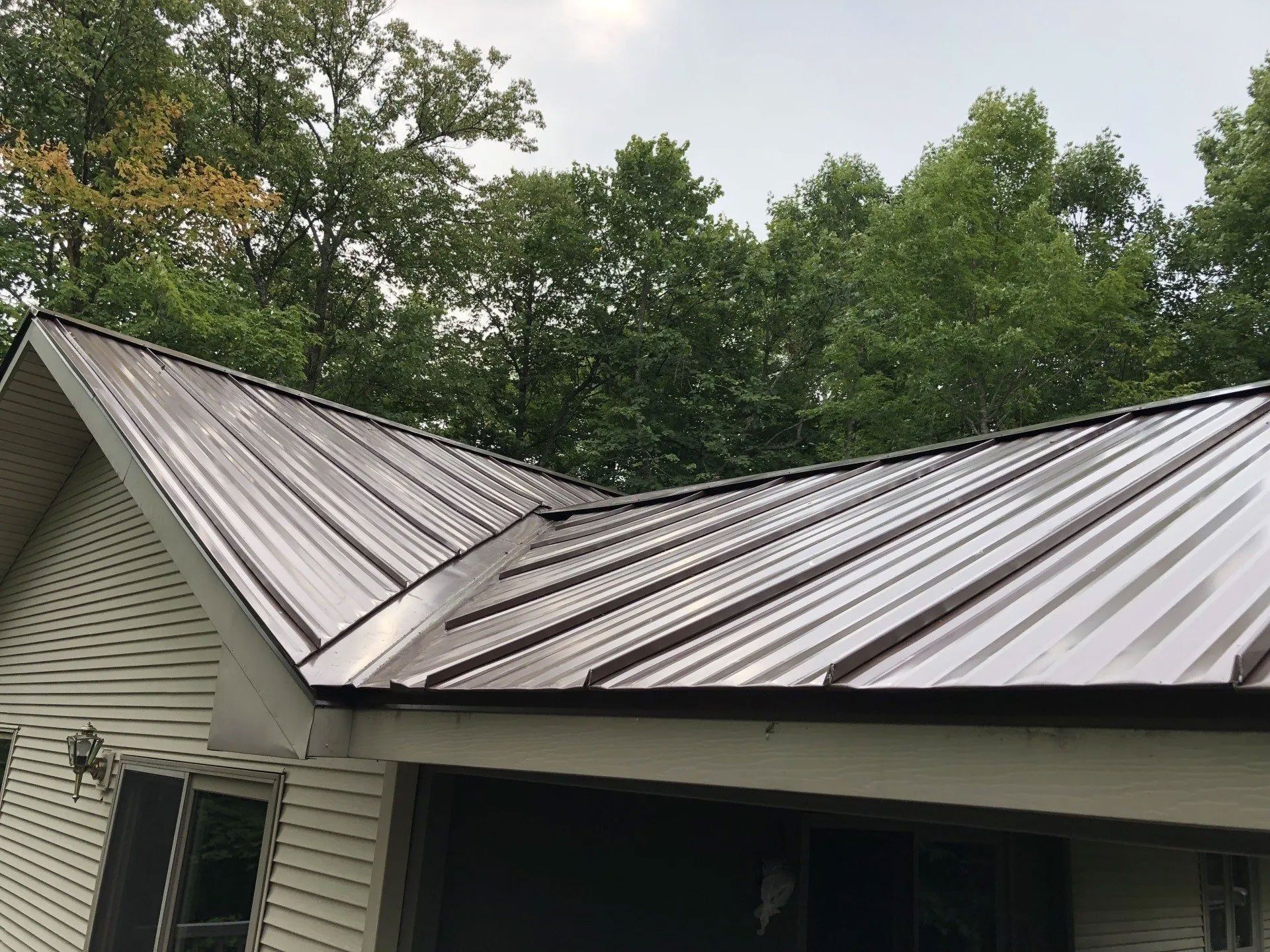 Metal roof on a white house with a damaged seam, surrounded by trees under an overcast sky