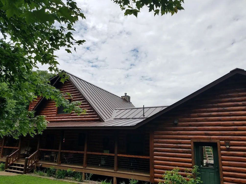 Log cabin with brown log siding and a metal roof, nestled among green trees under a cloudy sky