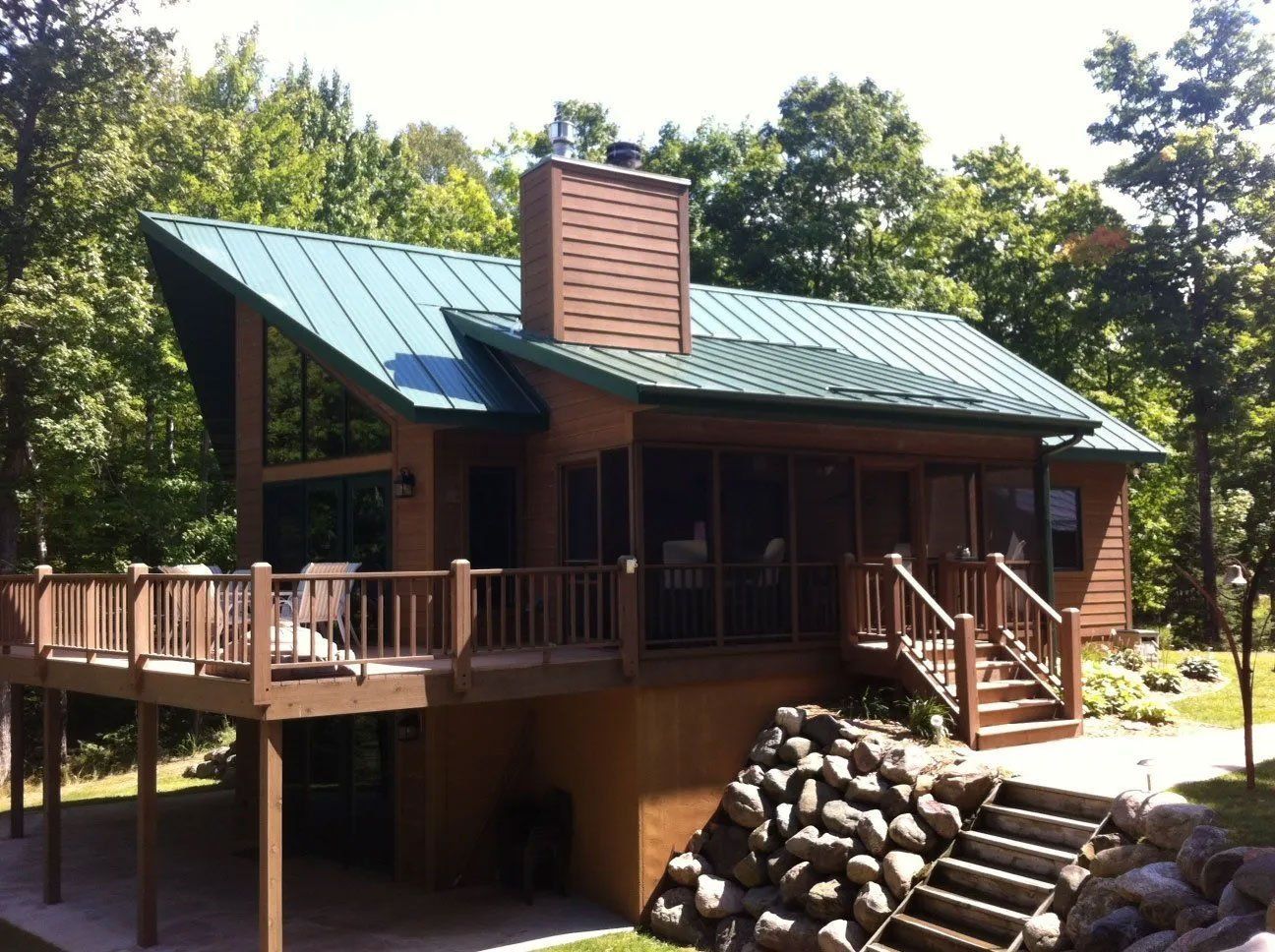 Wooden cabin with green roof and wraparound deck in a wooded area, with a stone retaining wall in front