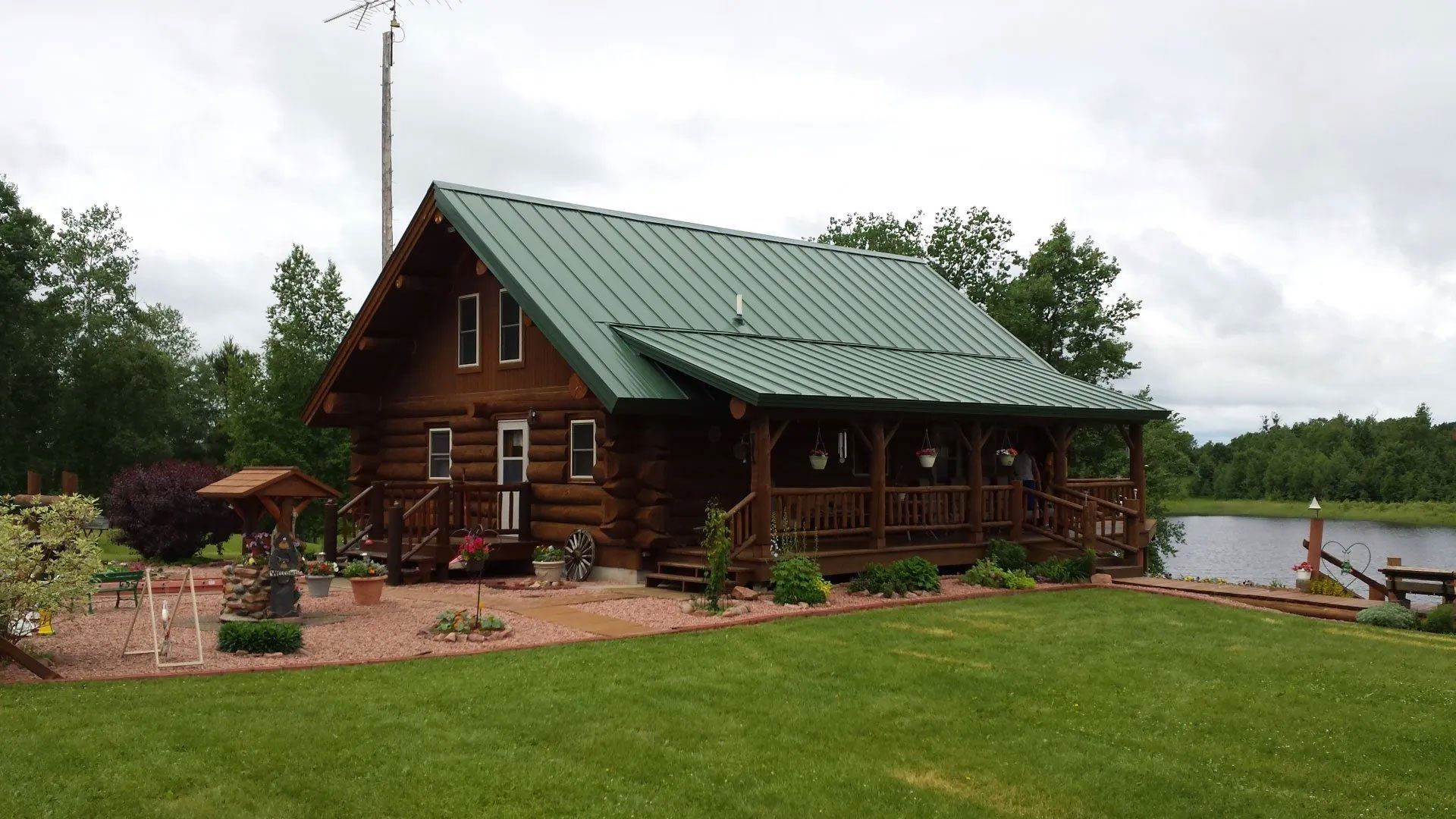 Rustic log cabin with green metal roof beside a lake, surrounded by trees and a grassy lawn.
