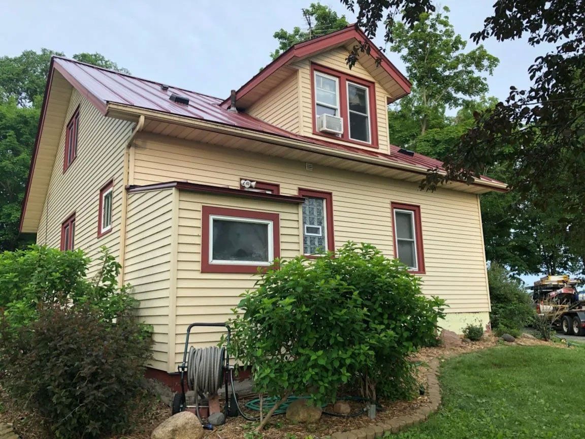 Yellow house with red trim, dormer windows, and shrubs in a grassy yard