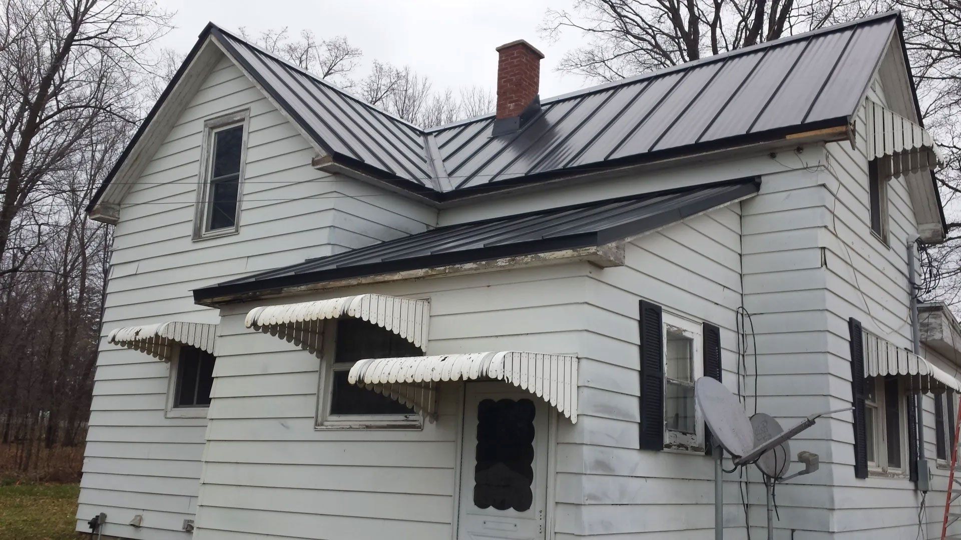 Dilapidated white house with sagging awnings and a metal roof in a wooded area