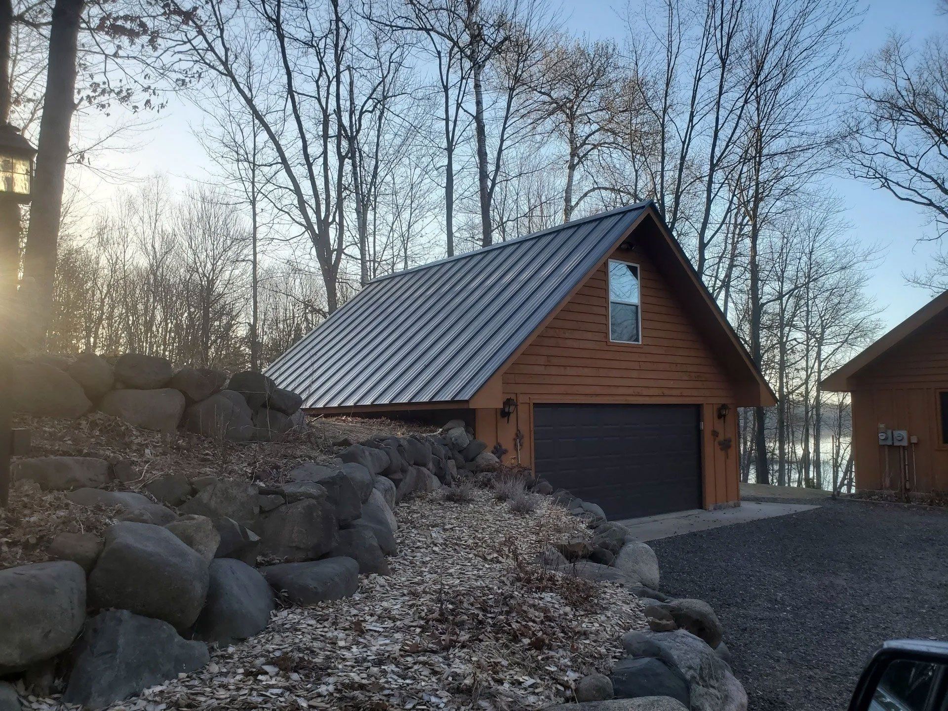 Wooden cabin with a metal roof beside a rocky driveway in a wooded yard at sunset
