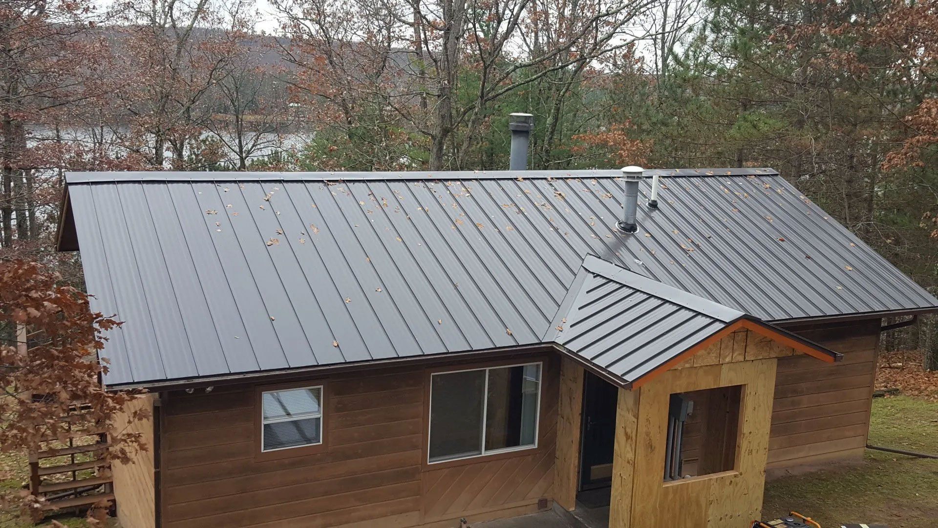 Small brown wooden house with a gray metal roof and a covered front entry, seen in a wooded area.