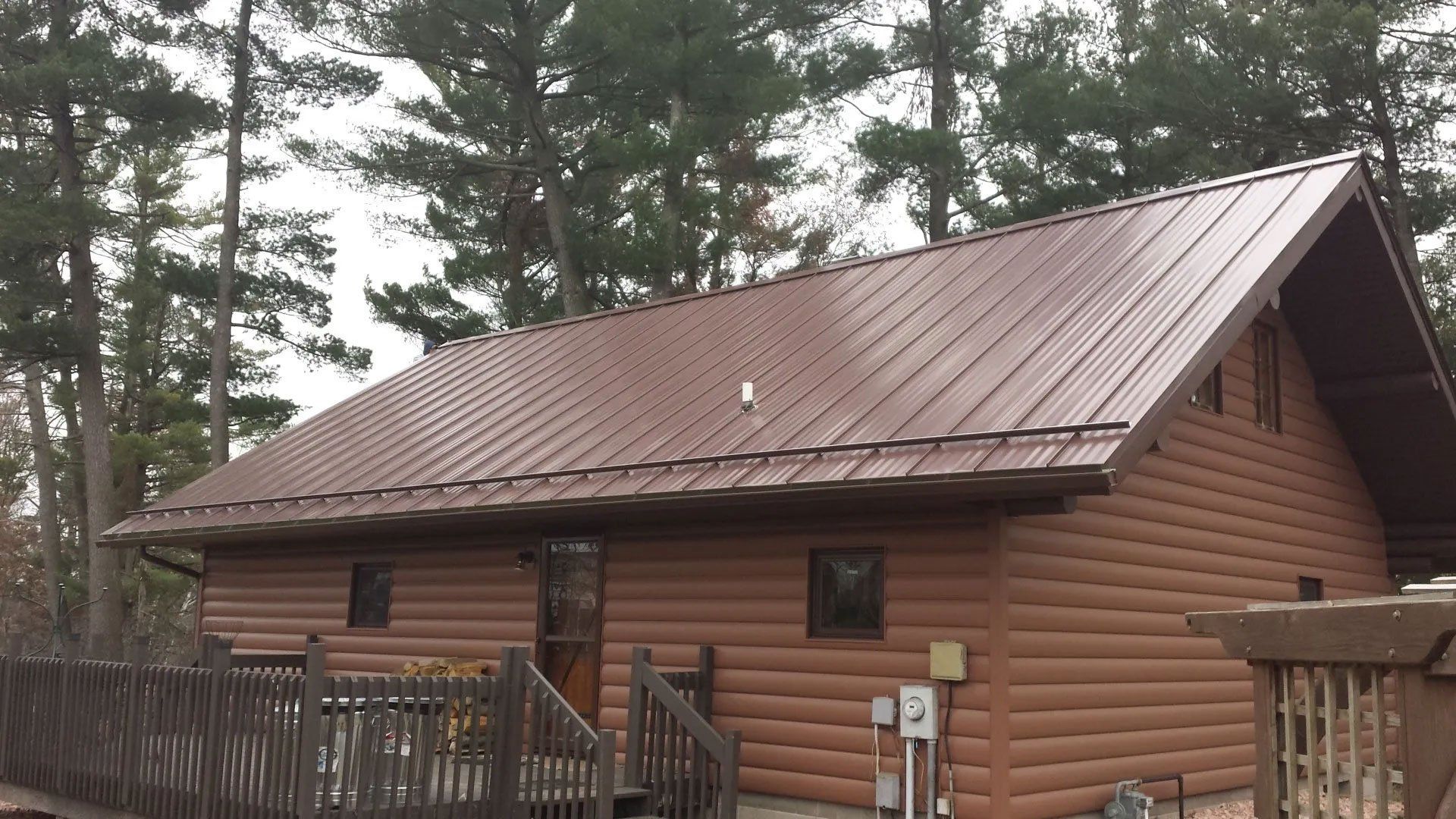 Wooden cabin with a brown metal roof behind a fenced yard and tall pine trees