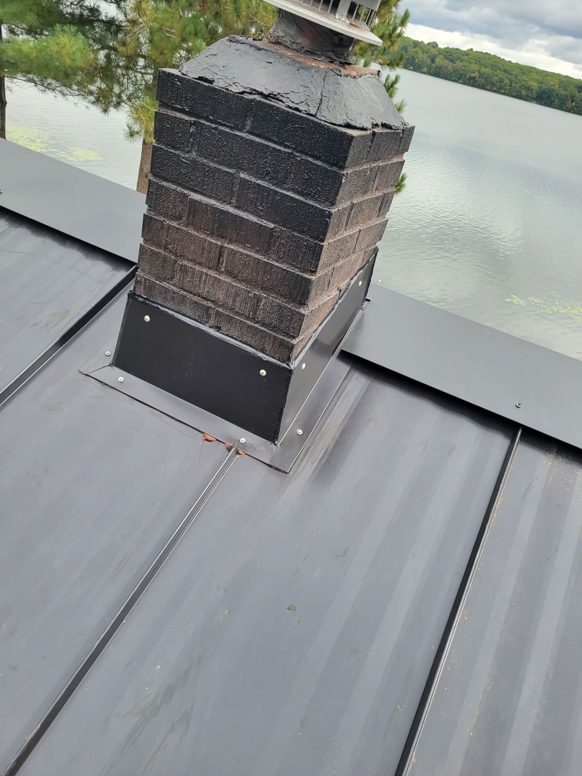 Rooftop vent chimney with dark flashing on a gray metal roof beside water