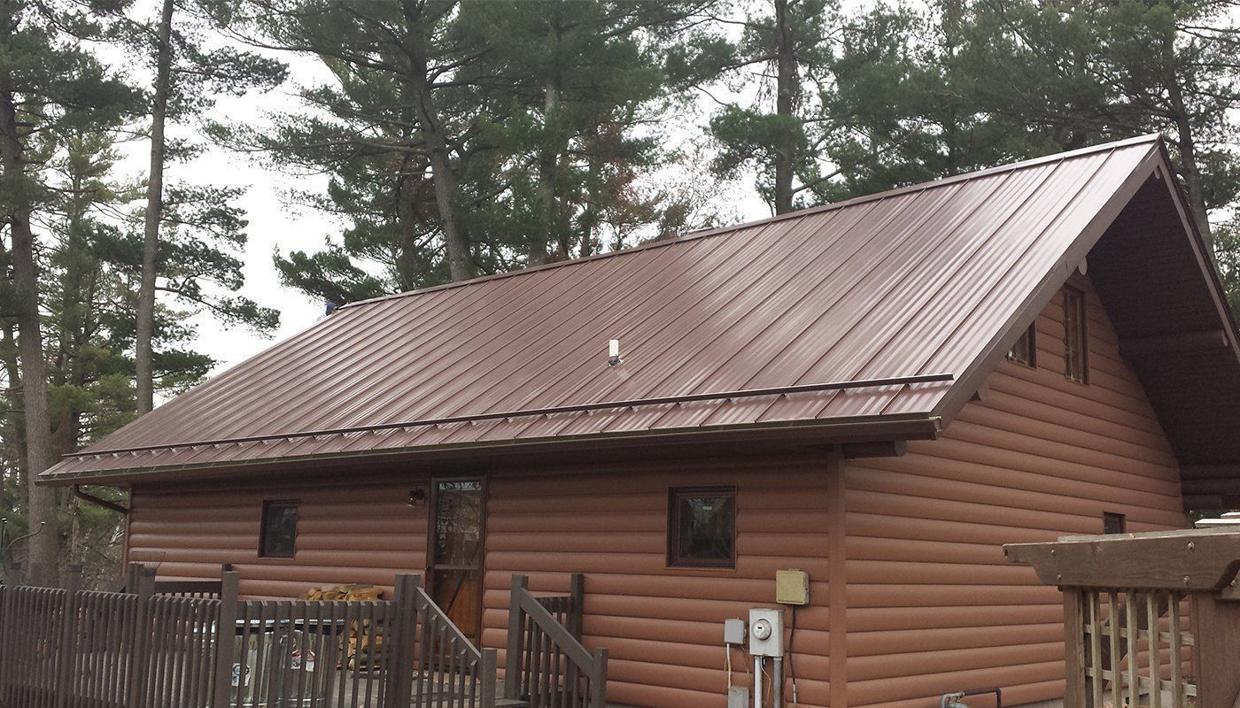 Wooden cabin with a brown metal roof amid tall pine trees and a fenced porch