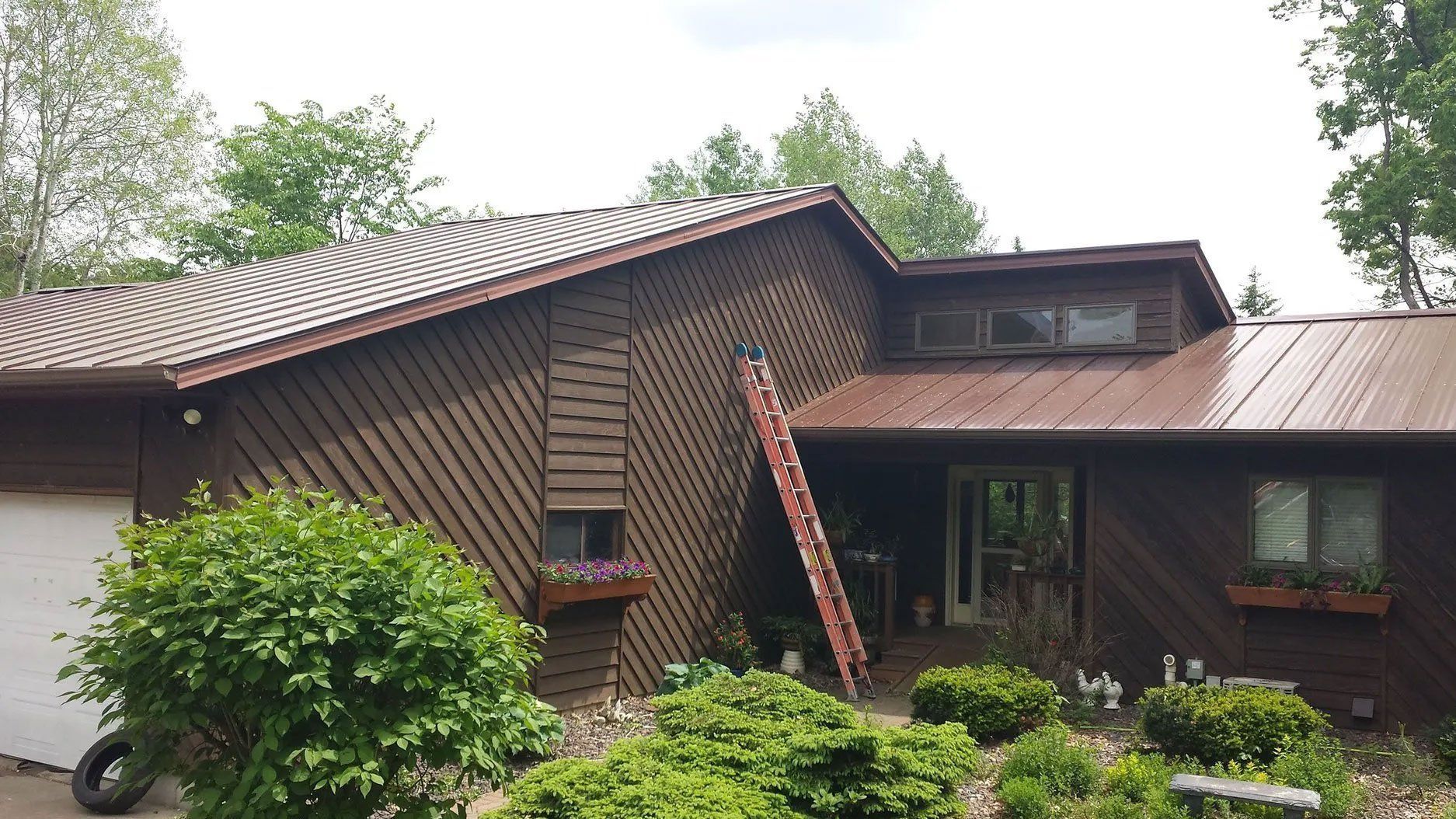 Rustic brown wooden house with a red-tiled roof, surrounded by green shrubs and trees, with a ladder leaning against it.