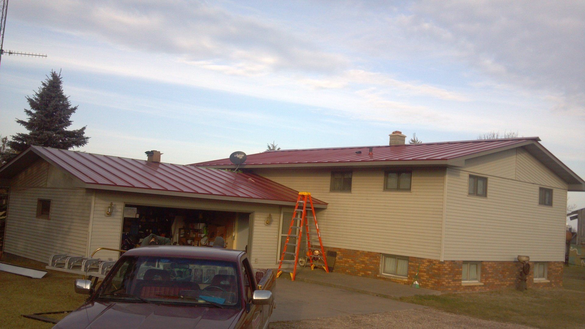 Two-story house with a red roof, ladders, and a car parked in the driveway under a cloudy sky