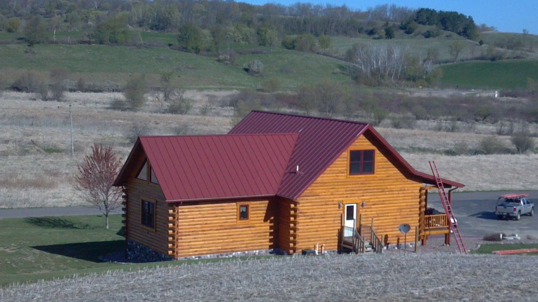Red-roofed wooden cabin in a grassy rural field with hills in the background