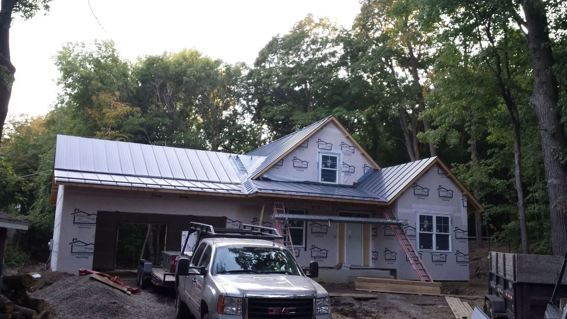 House under construction with metal roof, porch scaffolding, and a truck parked in front, surrounded by trees