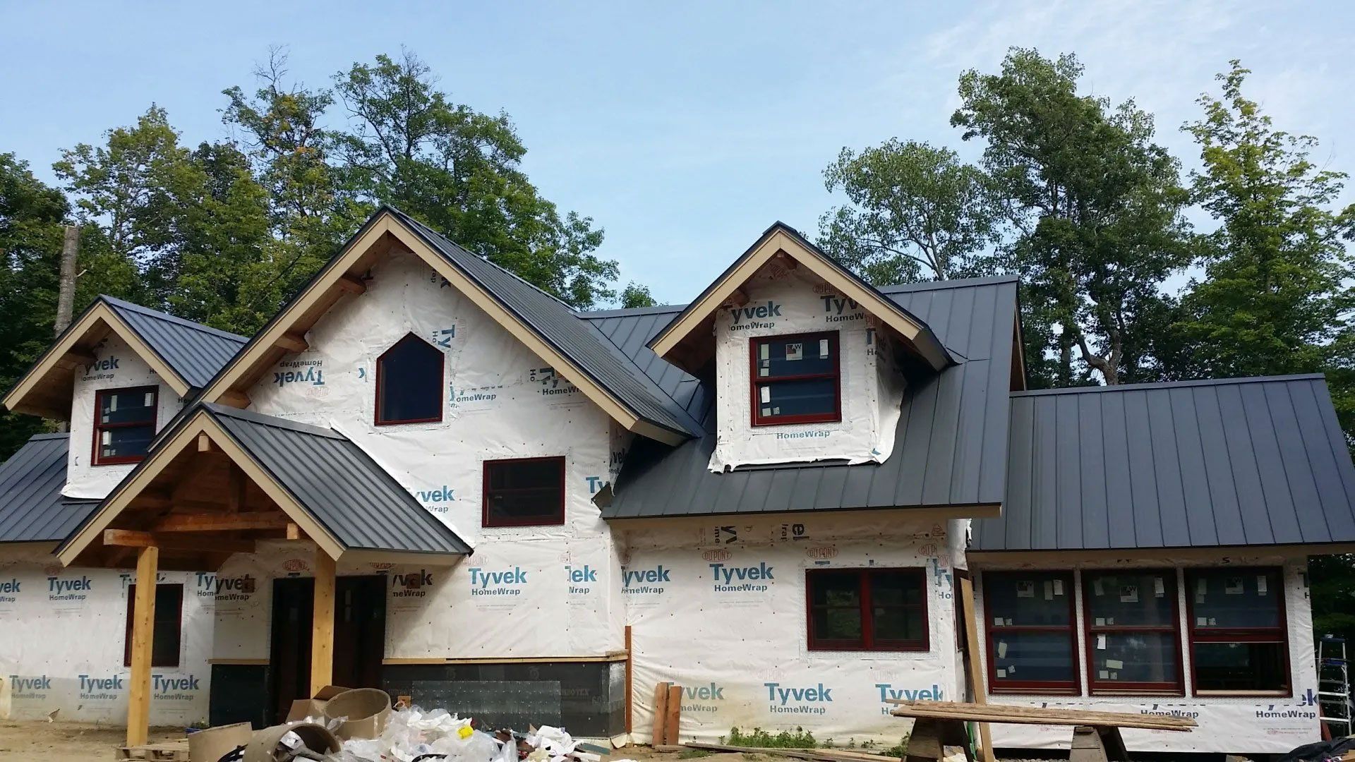 House under construction with white siding, black roof, and a covered front porch, surrounded by trees.