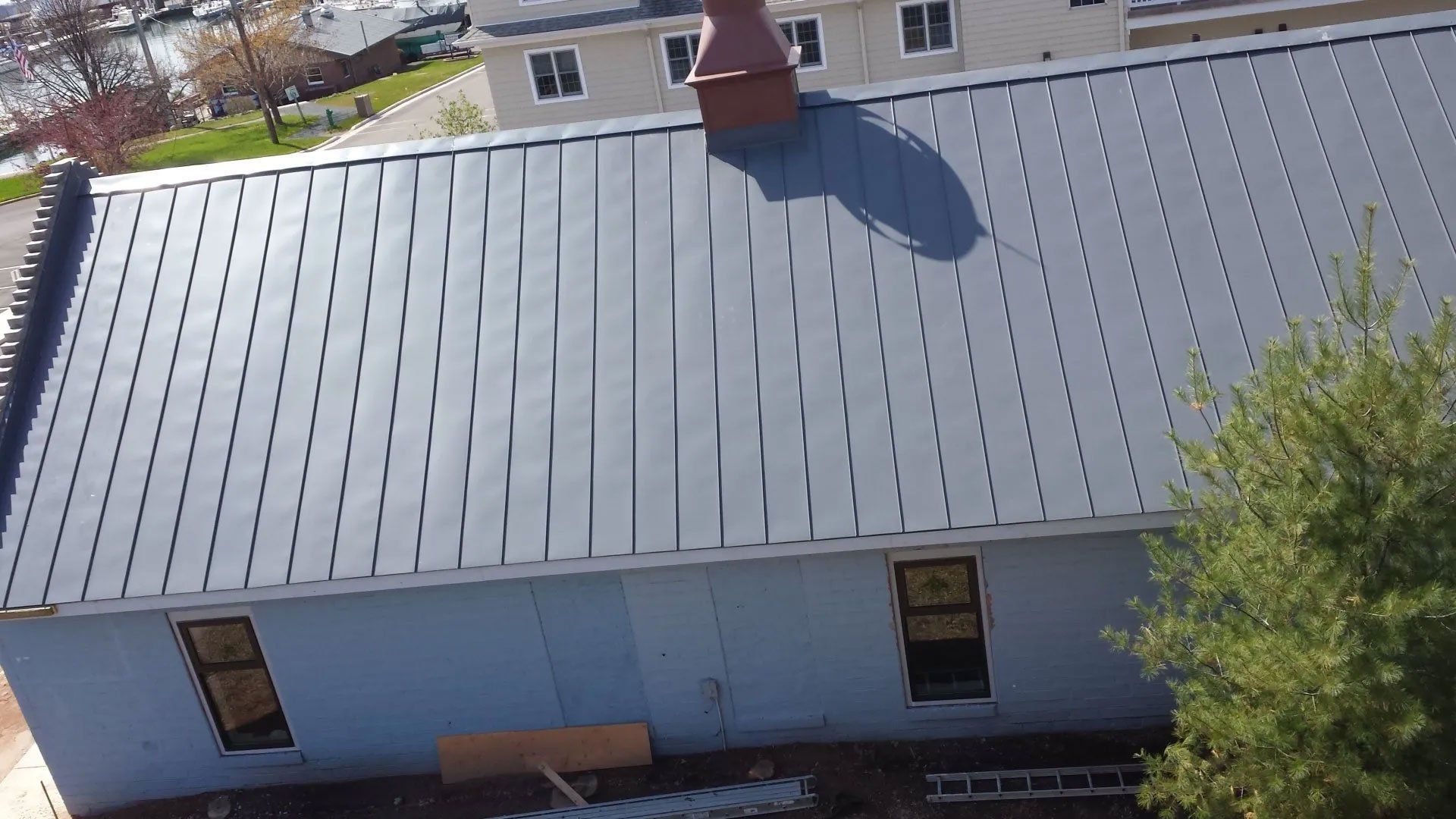 Blue house with a gray metal roof and chimney, viewed from above with trees and snow at the edge