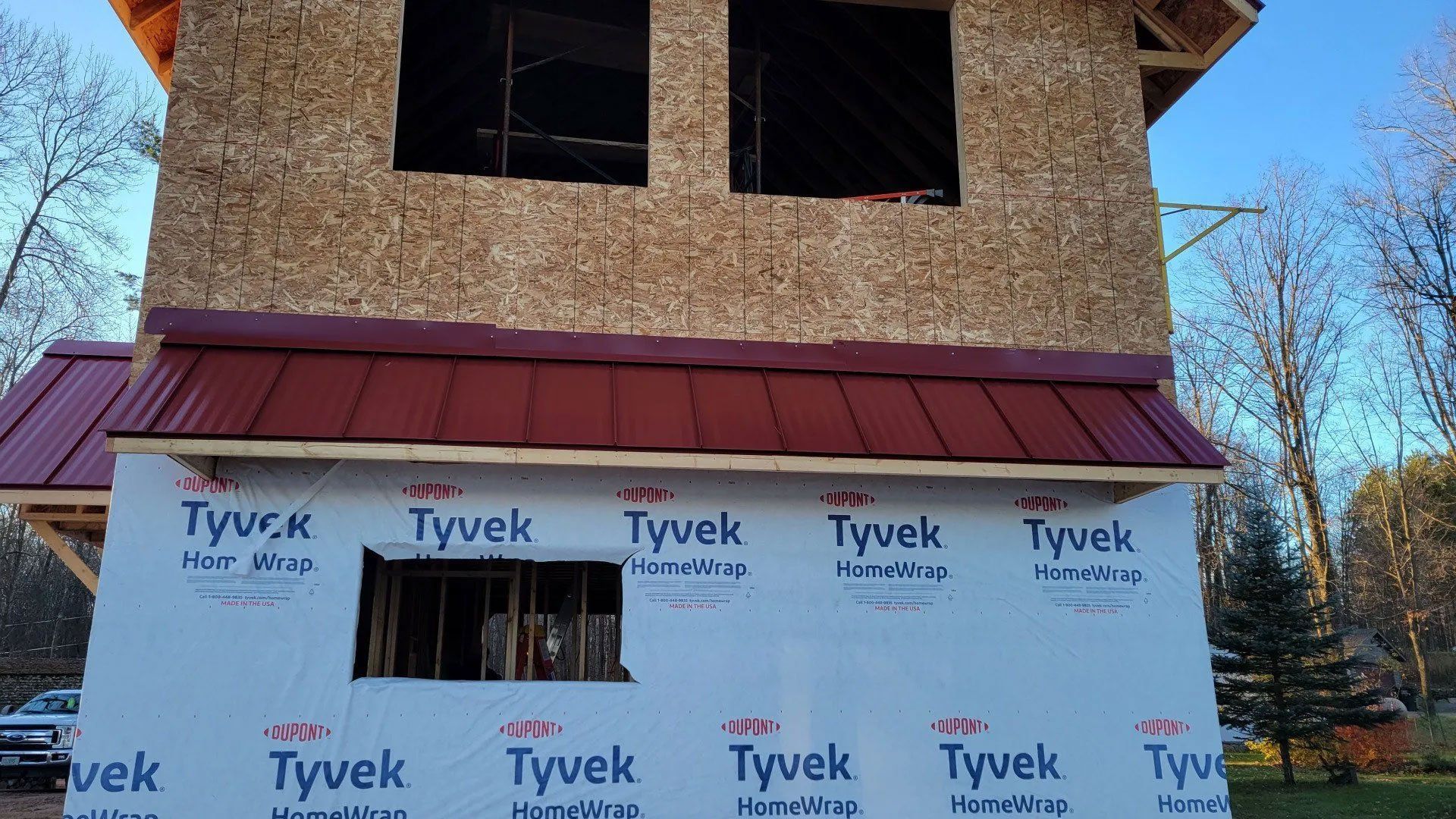 House under construction with Tyvek wrap, red metal roof, and open window and door openings.