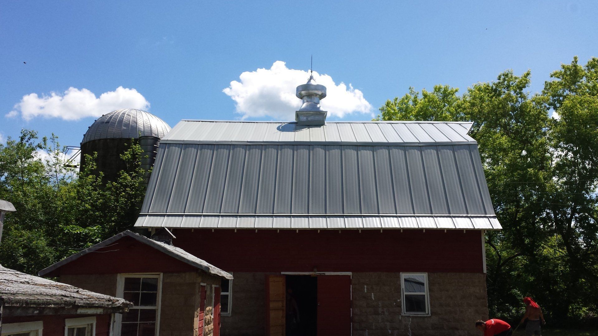 Small red brick building with a gray metal roof and cupola under a blue sky with trees and clouds