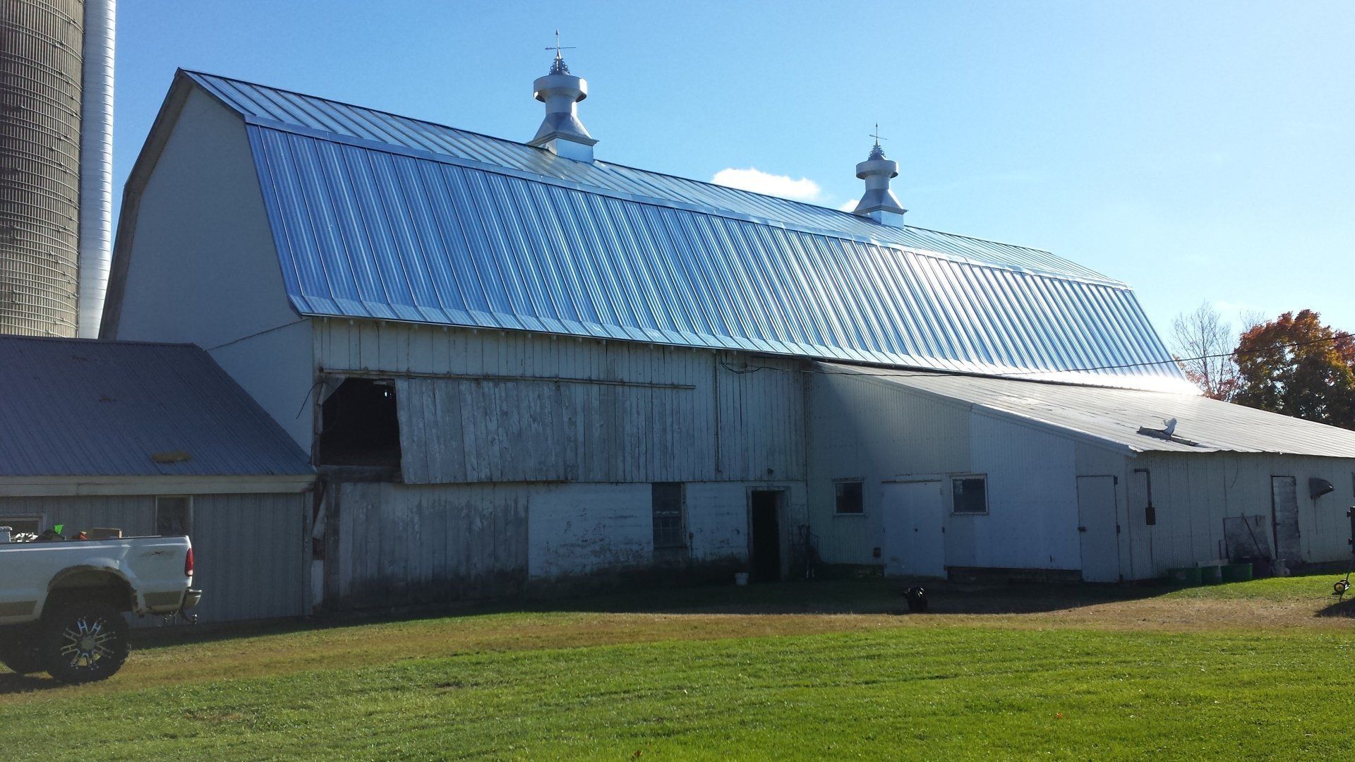 Large white barn with a blue metal roof and cupolas on a grassy field under a clear sky