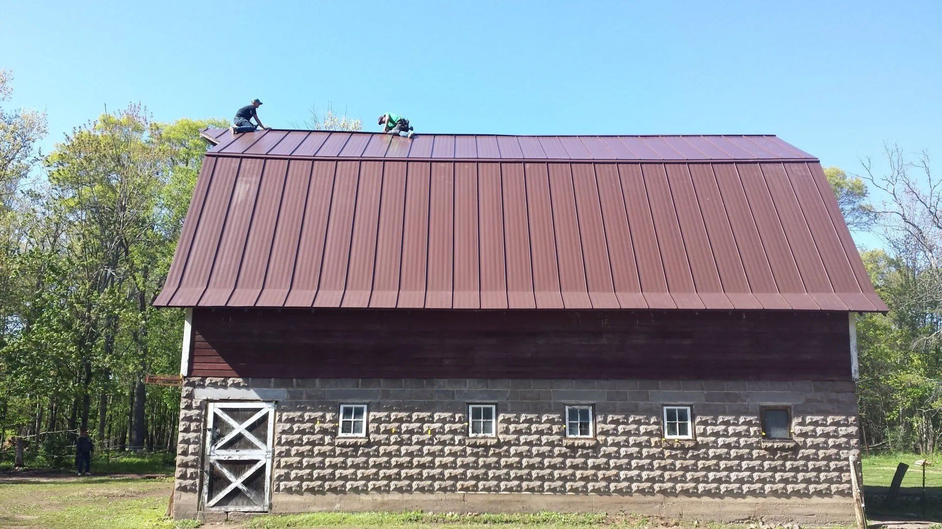 Red barn with a brown roof and stone base under a blue sky, with trees in the background