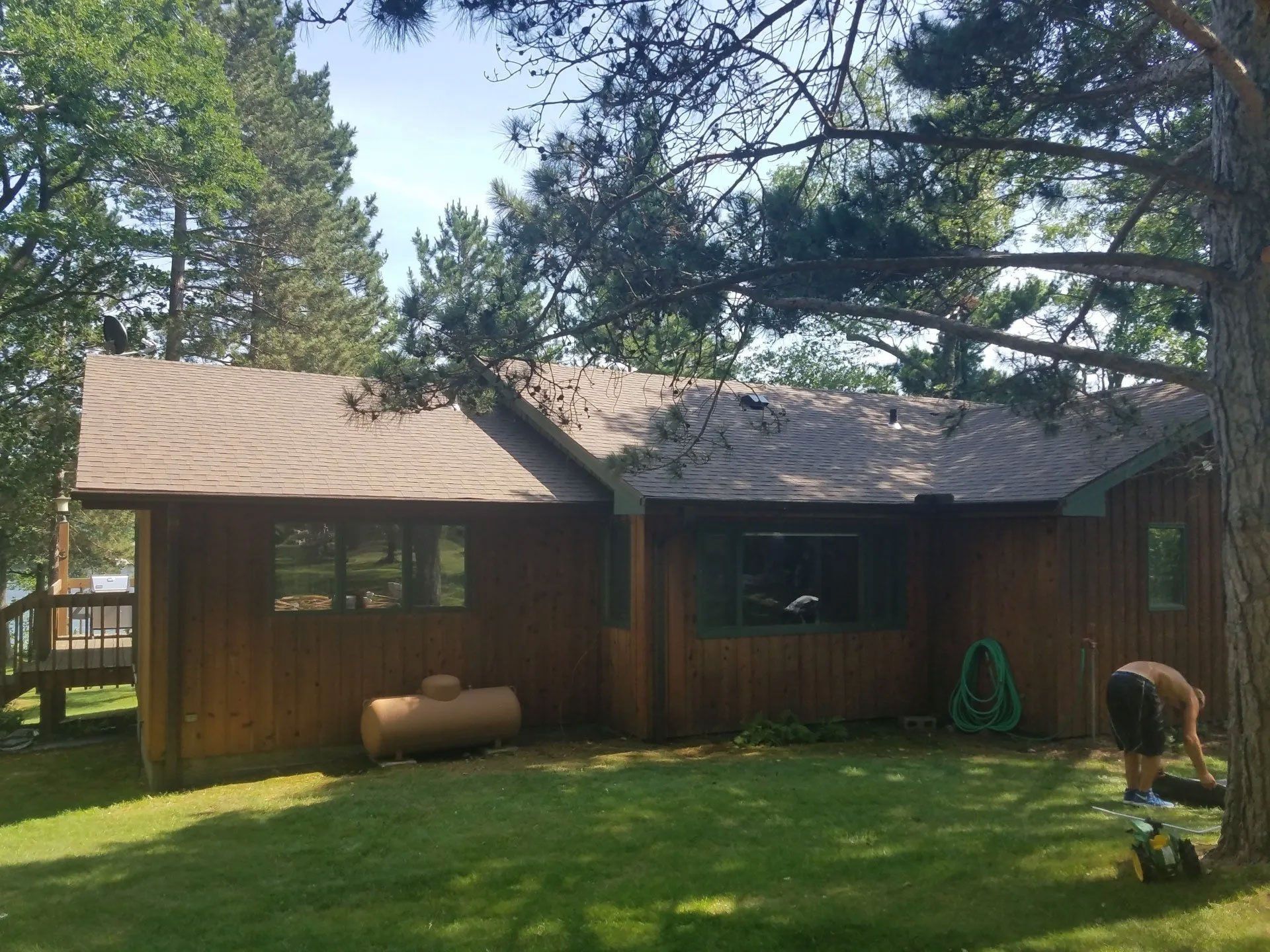Wooden cabin with a green lawn, tall trees, and a person watering plants on the right.