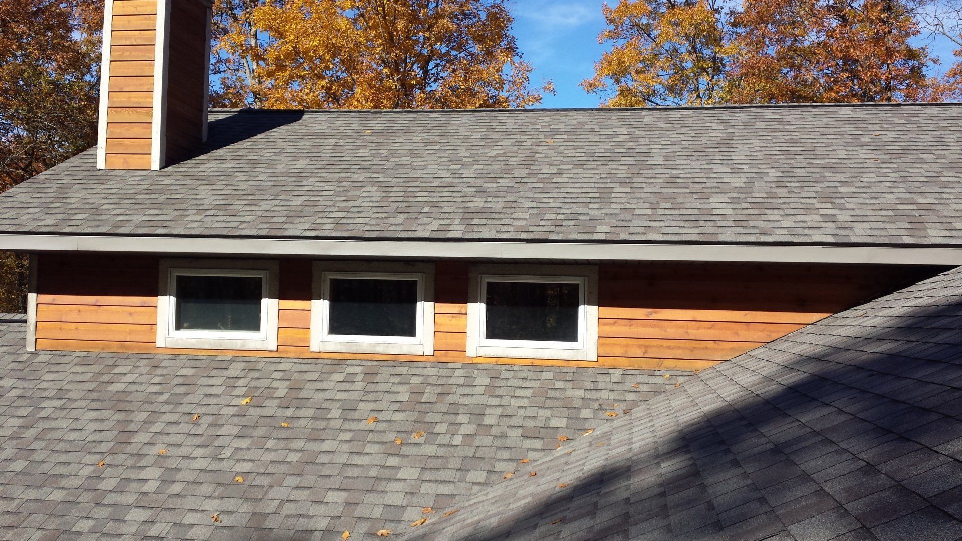 House roof with three small windows, brick chimney, and autumn trees in the background