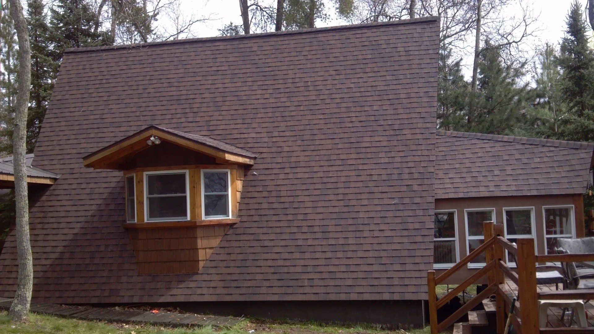 Brown cabin with steep shingled roof, small dormer window, and wooden deck in a wooded setting