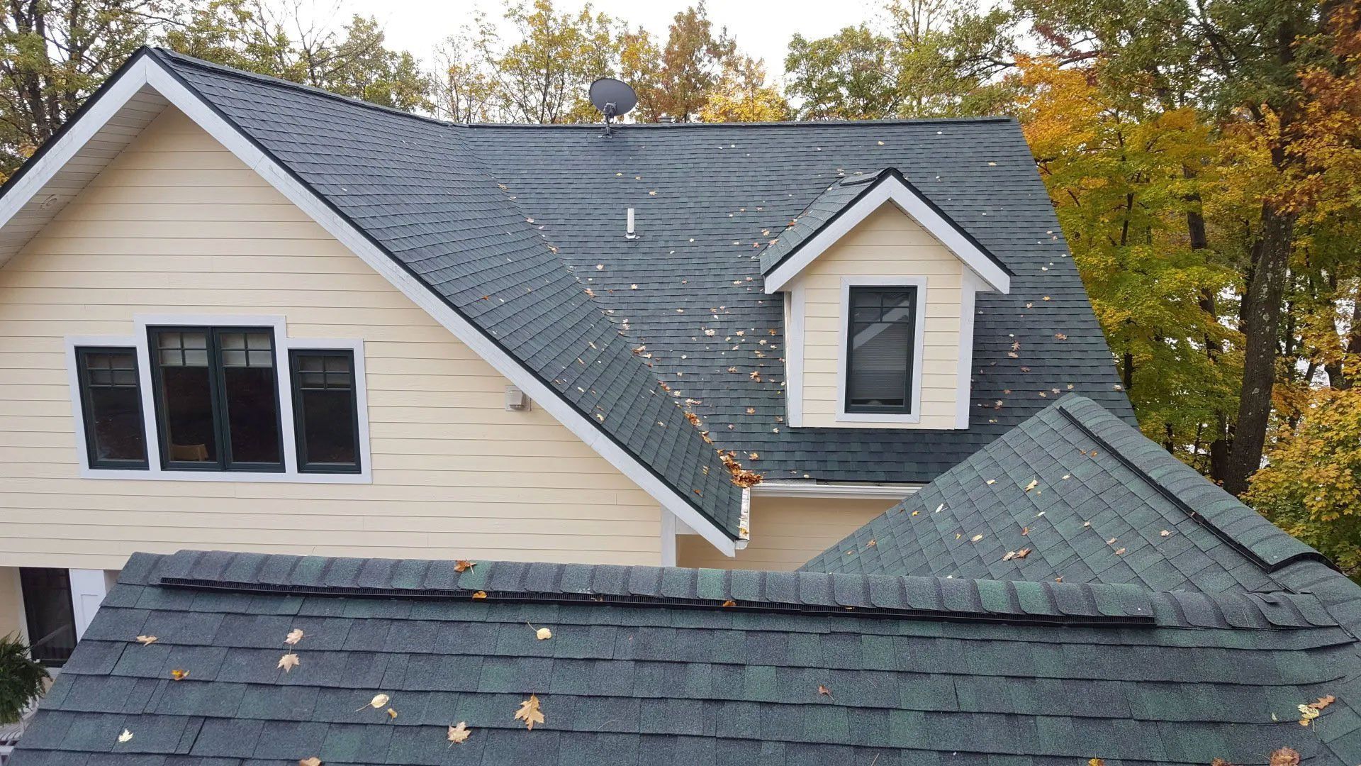 Roof with gray shingles and dormer windows, seen from above with autumn trees in the background
