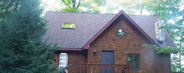 Brown wooden house with a steep roof, front deck, and surrounding trees
