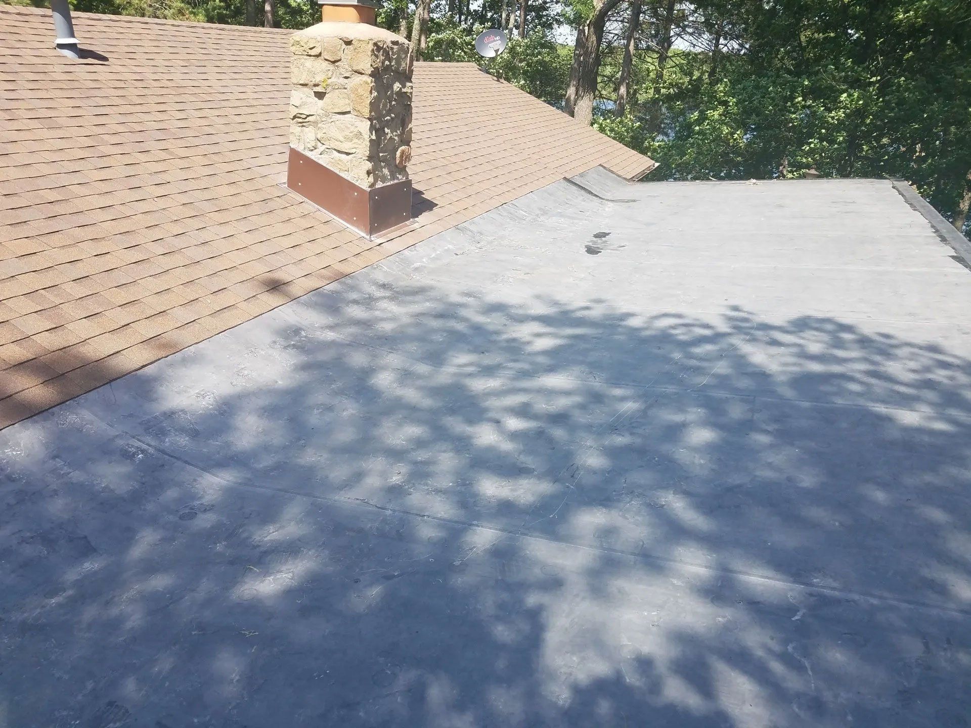 Rooftop with shingles, stone chimney, and gray flat surface under tree shadows